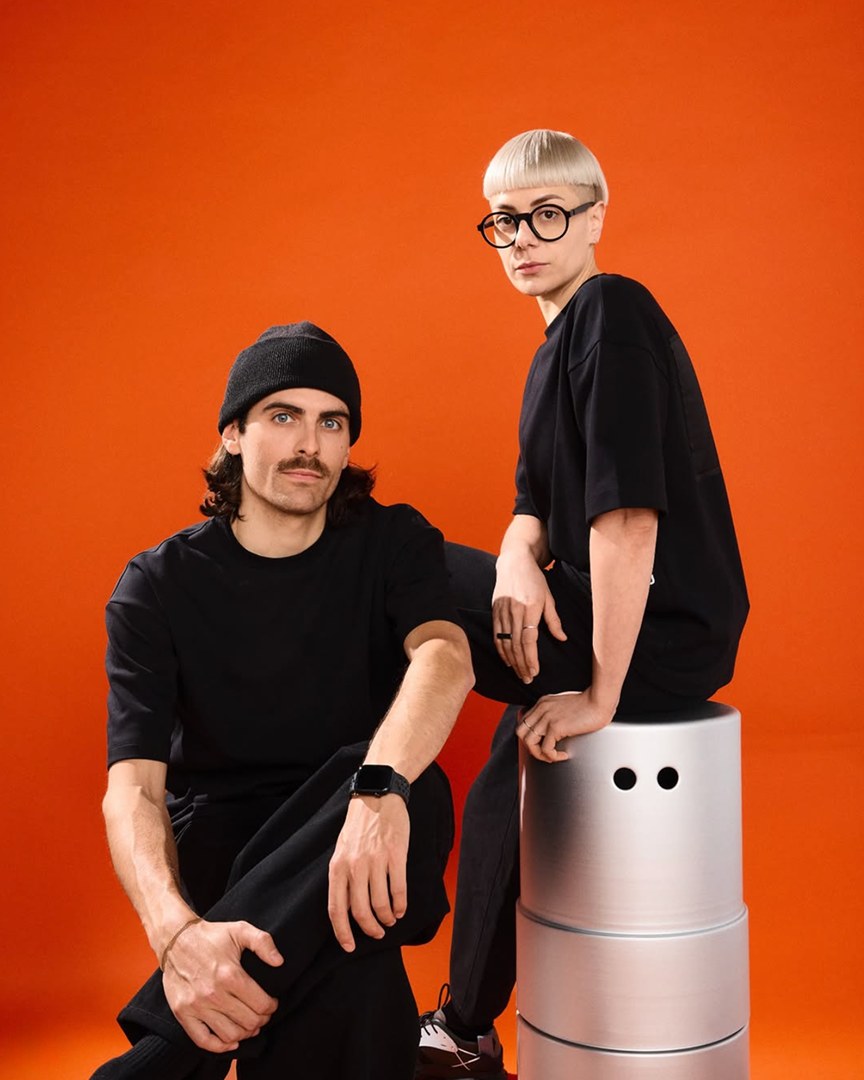 Odd Universe co-founders seated on a bold orange backdrop beside a cream Peepers cylinder product