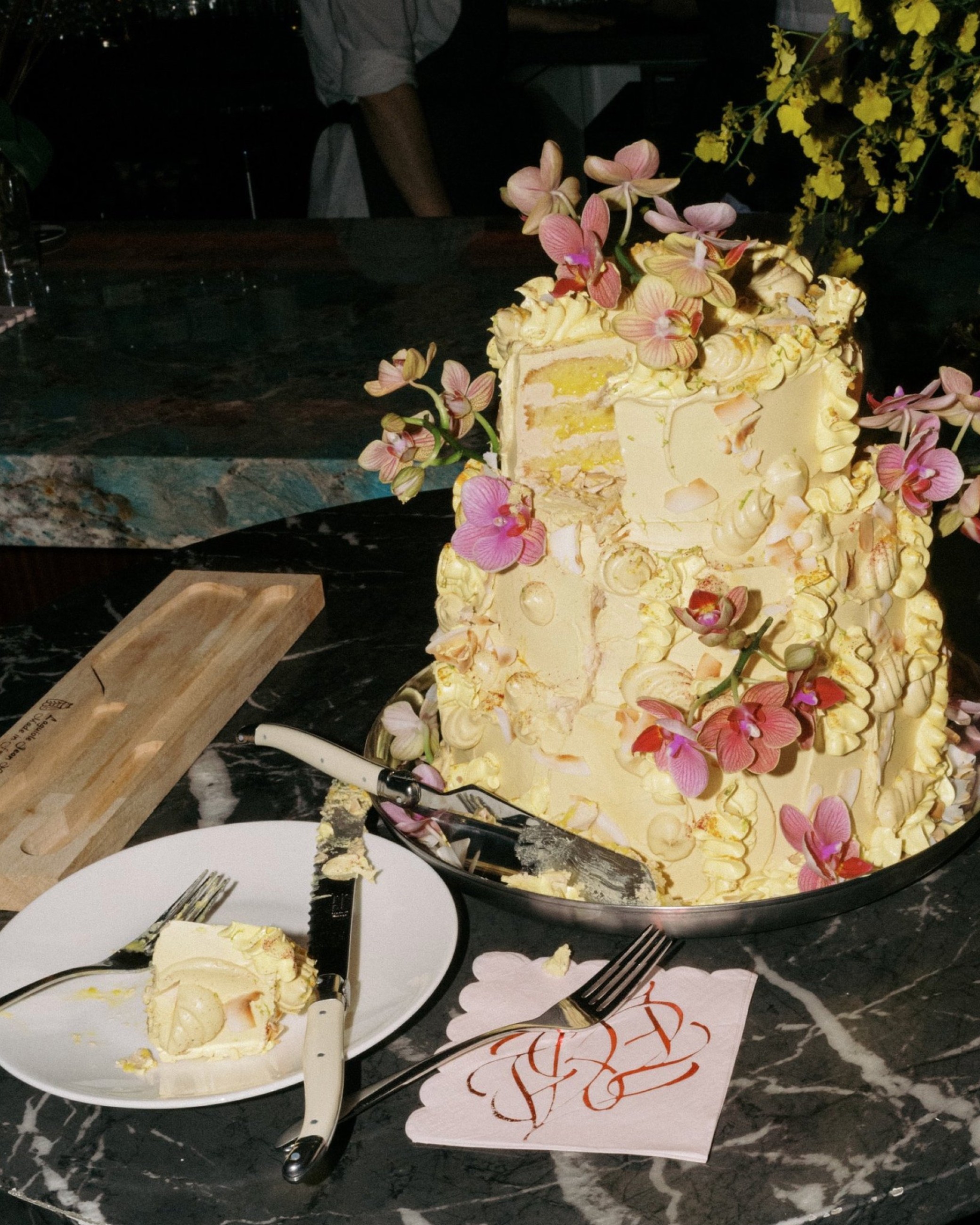 Atelier Amarise buttercream wedding cake with pink orchids, a cut slice and monogrammed napkin on black marble