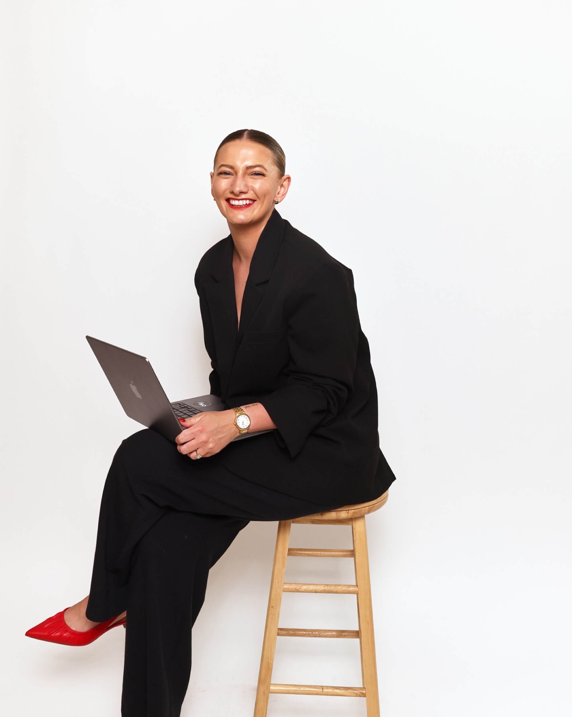 Alice Benham on a wooden stool with a laptop, wearing a black suit and red heels, brand photoshoot