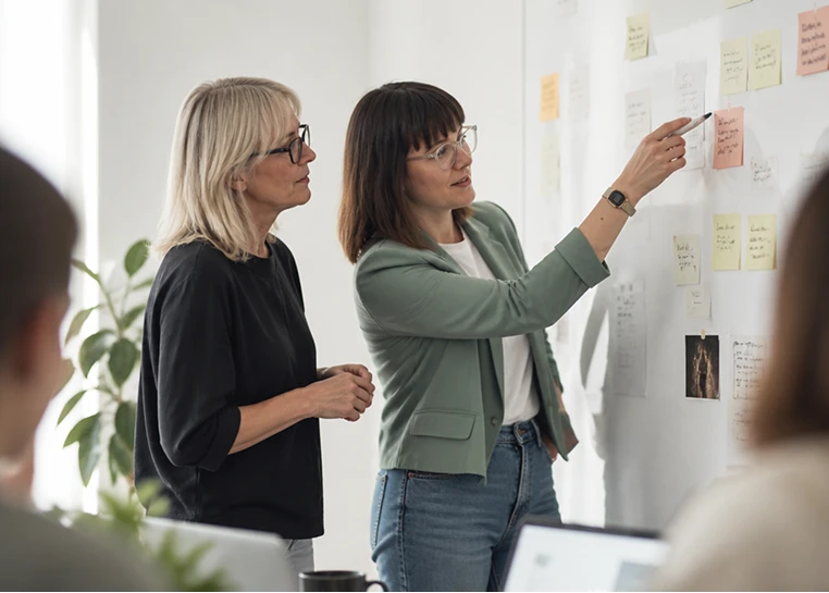 Deux femmes regardent une autre femme qui pointe des notes sur un mur lors d’une réunion de travail.