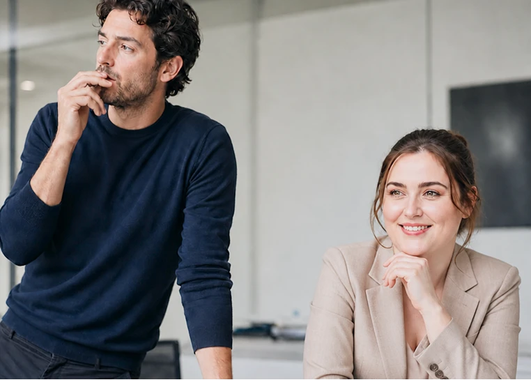 Un homme pensif en chandail bleu et une femme souriante en veston beige dans un bureau moderne.