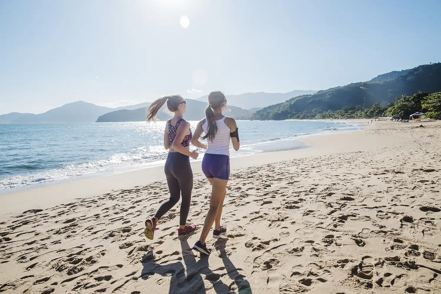 Deux femmes courant sur plage ensoleillée, paysage côtier, activité sportive en extérieur.