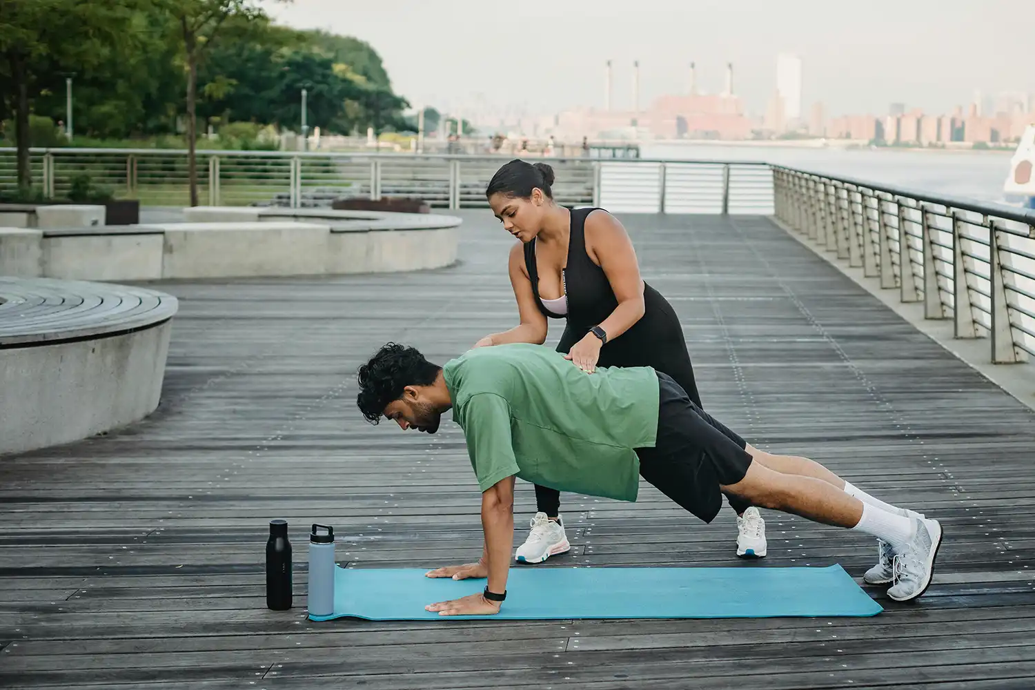Deux personnes font un exercice de gainage en extérieur sur tapis de sport près d’un plan d’eau, idéal pour séance fitness en plein air.