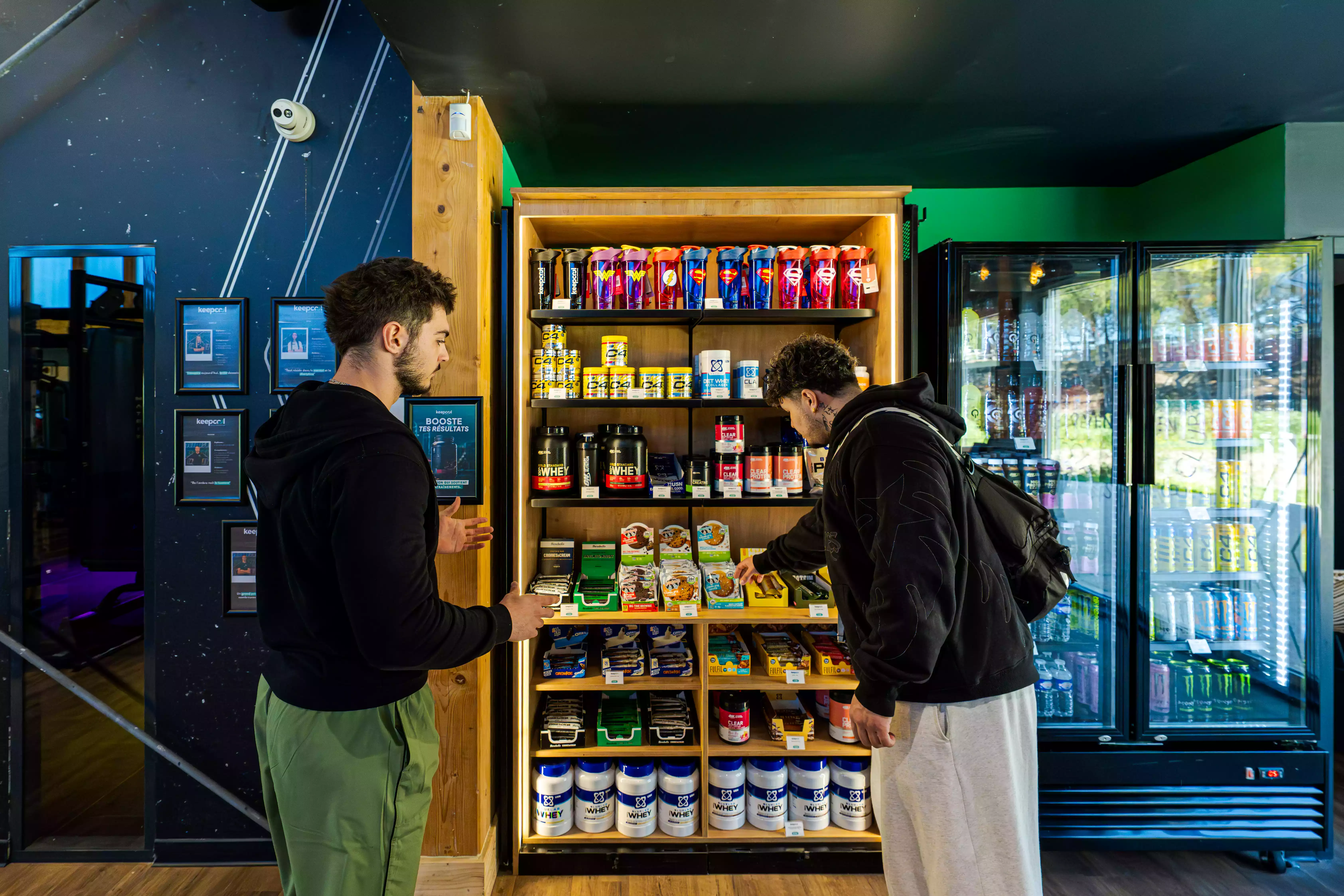Deux personnes choisissent des boissons et compléments alimentaires dans un espace nutrition Keepcool, frigo et étagères remplis de produits healthy.