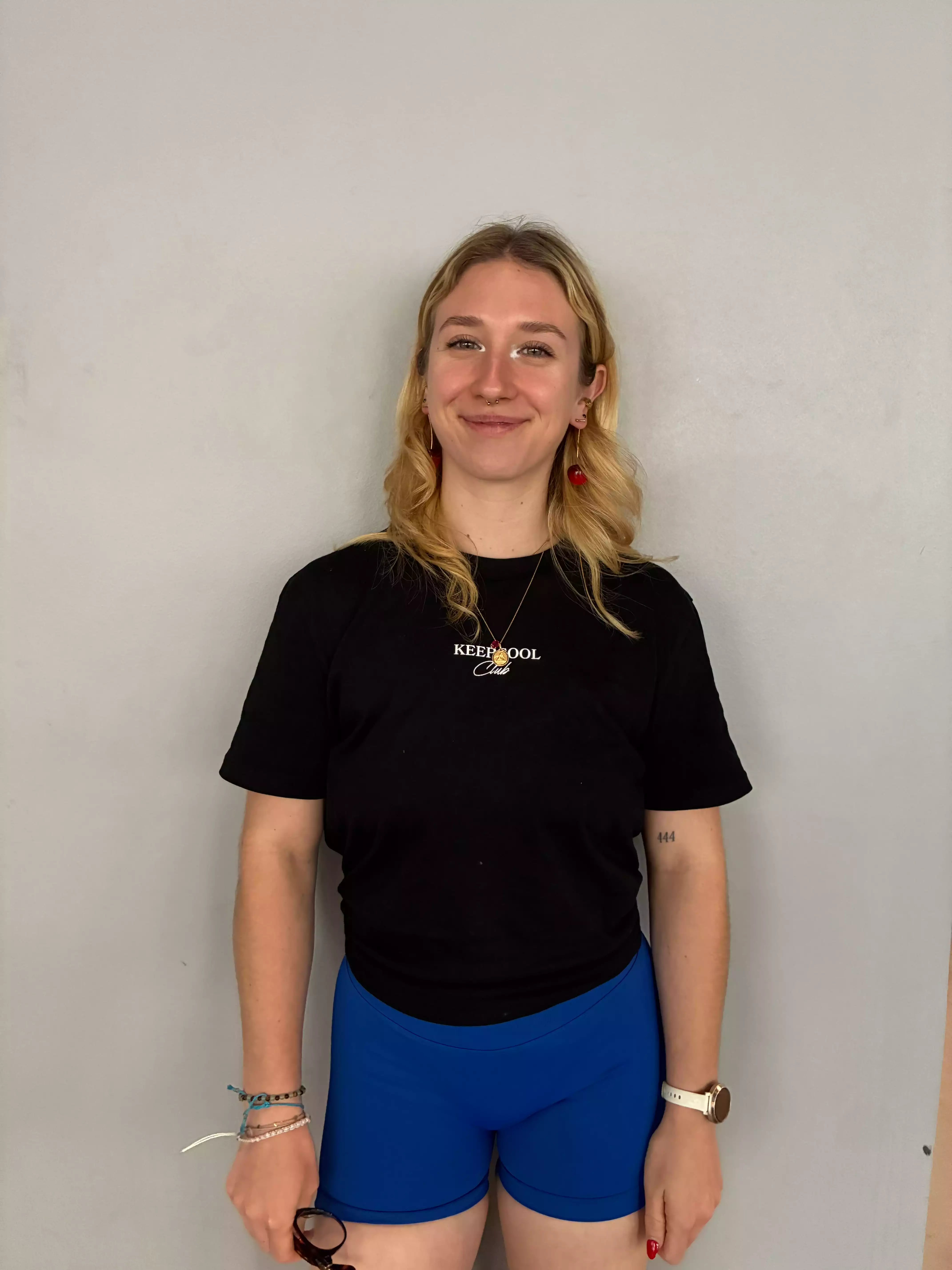 Femme souriante en tenue sportive Keepcool, short bleu et t-shirt noir, devant fond blanc.