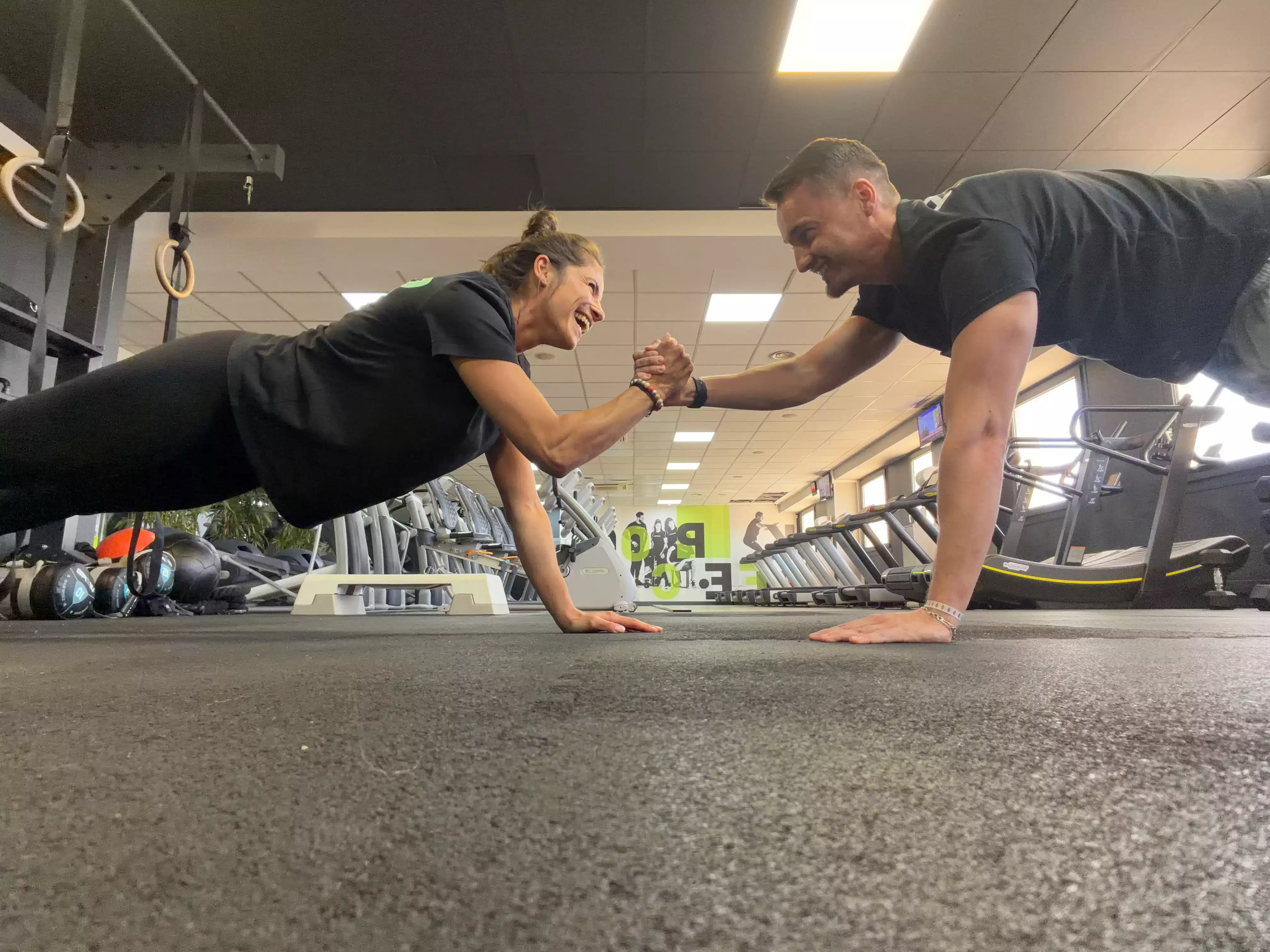 Deux athlètes en salle de sport réalisant un exercice de gainage au sol, main contre main, environnement gymnique moderne.