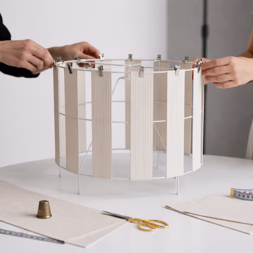 Two pairs of hands attaching beige fabric strips with metal clips to a round wire frame for lampshade making on a white table with scissors, thimble, and measuring tape.
