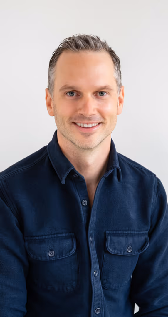 Smiling man with short hair wearing a dark blue button-up shirt against a plain background.