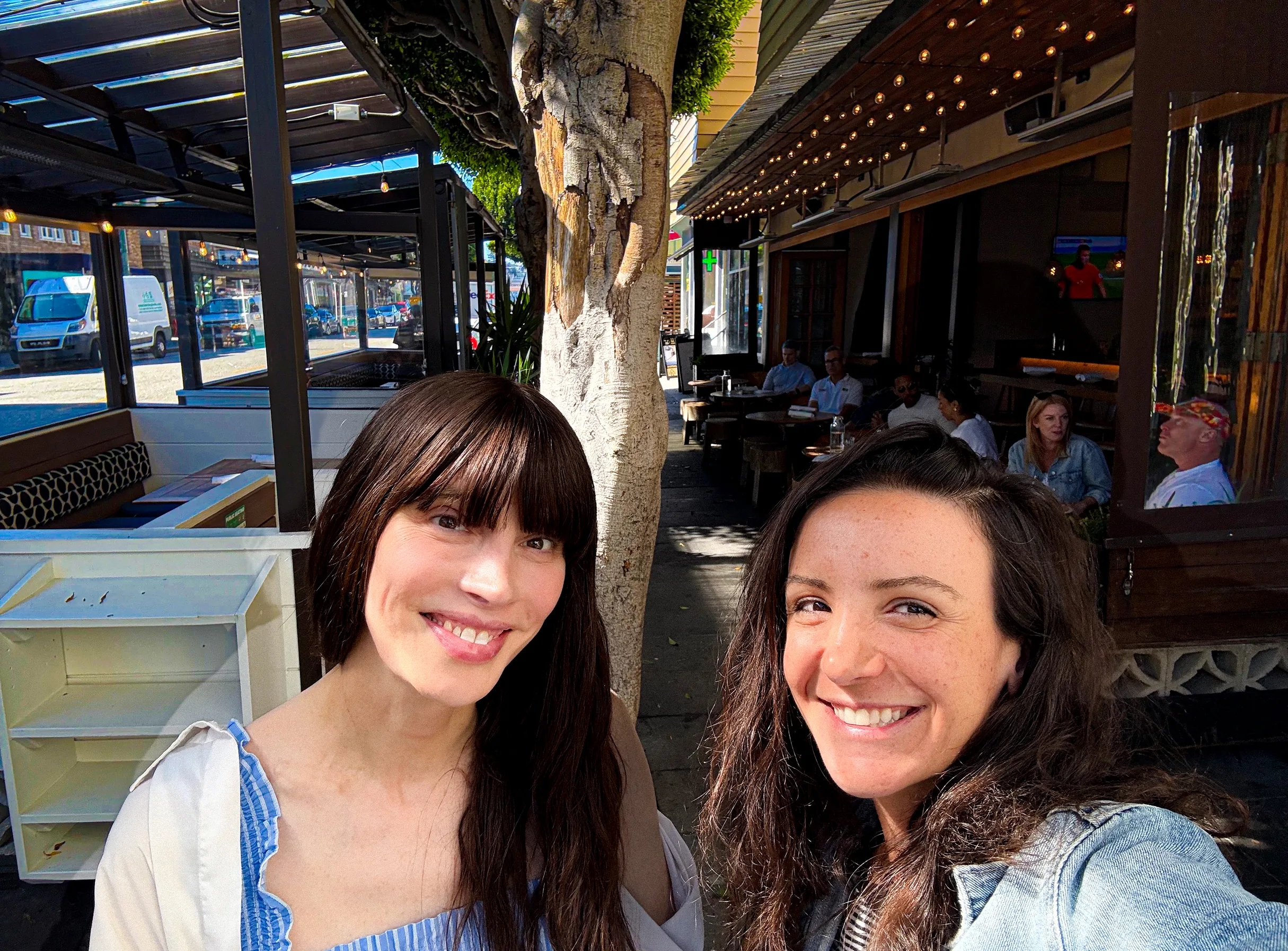 Two smiling women taking a selfie at an outdoor café with tables and people in the background.