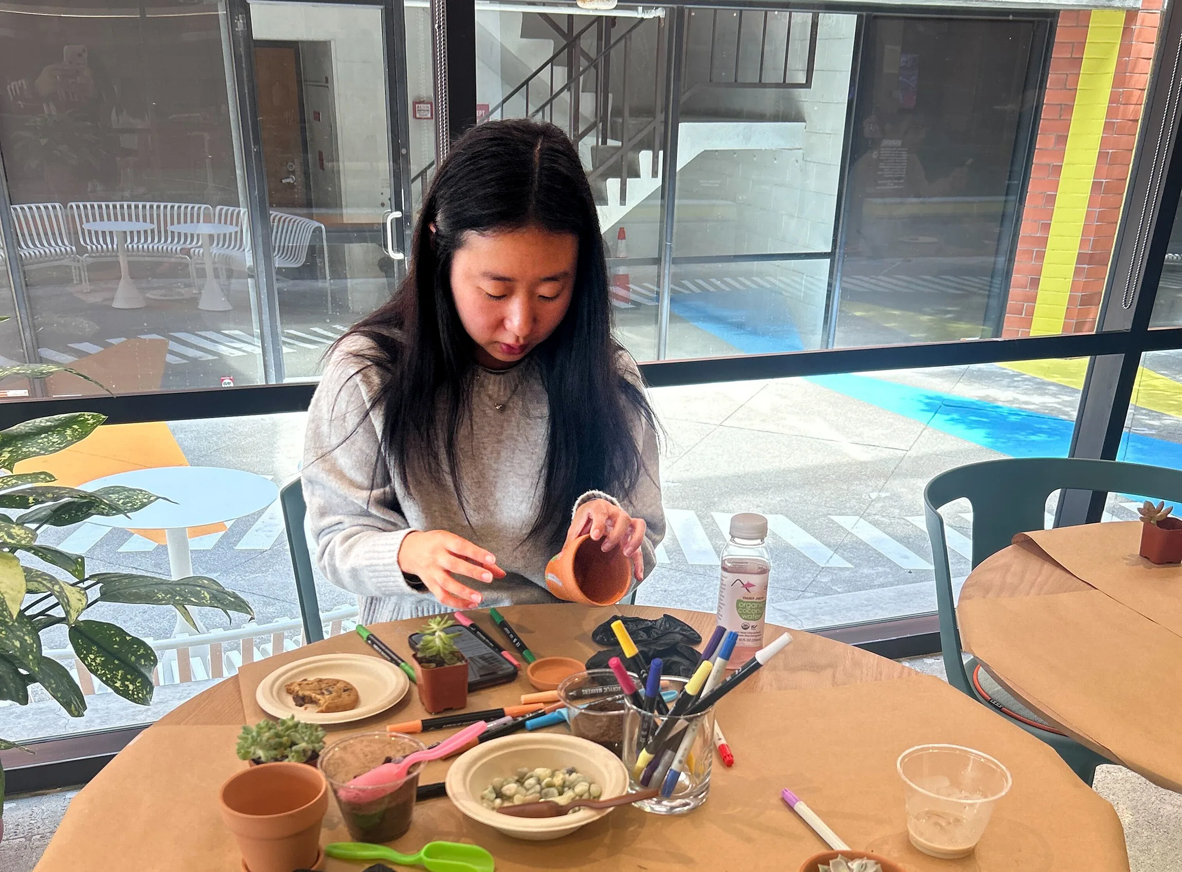 Person sitting at a table painting a small terracotta pot with markers and plants nearby.