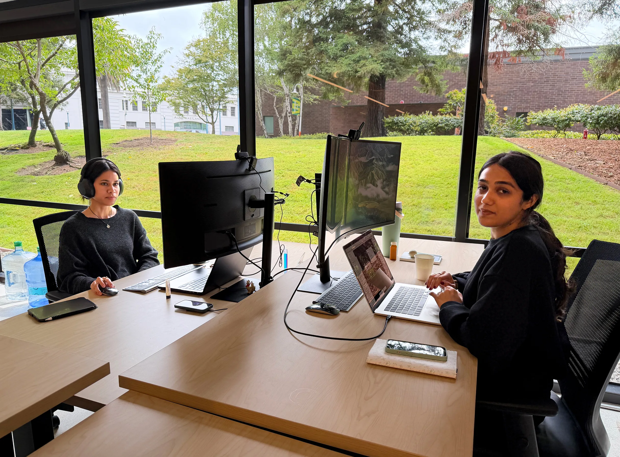Two women working at desks with computers in an office with large windows showing a grassy outdoors.
