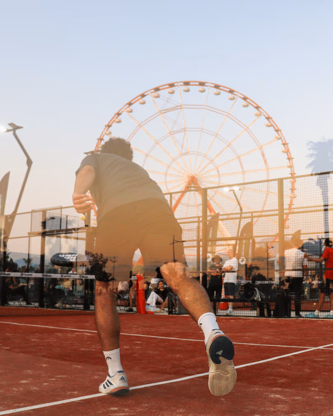A padel player on a red padel court, in reflection of the padel court glass there is an panoramic wheel.