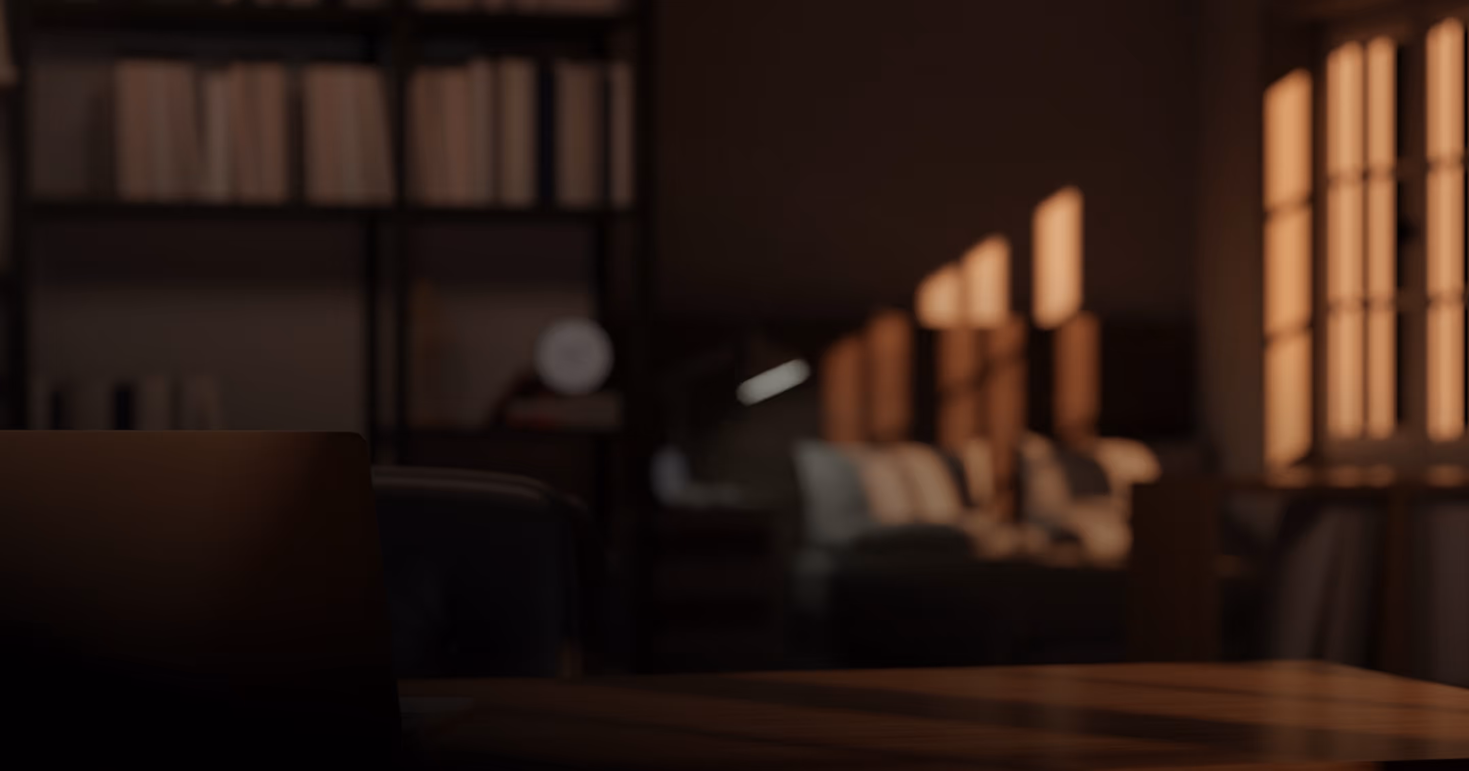 Laptop partially open on wooden desk with blurred bookshelf and warm window light in background.