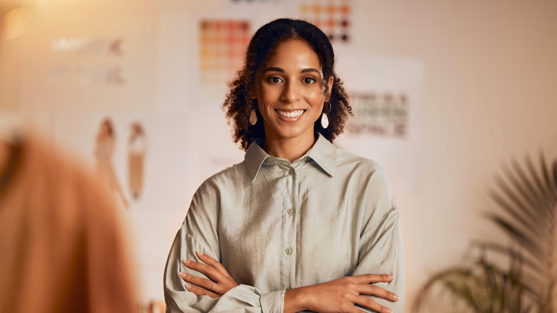Confident woman with curly hair smiling with arms crossed in a softly lit office space.