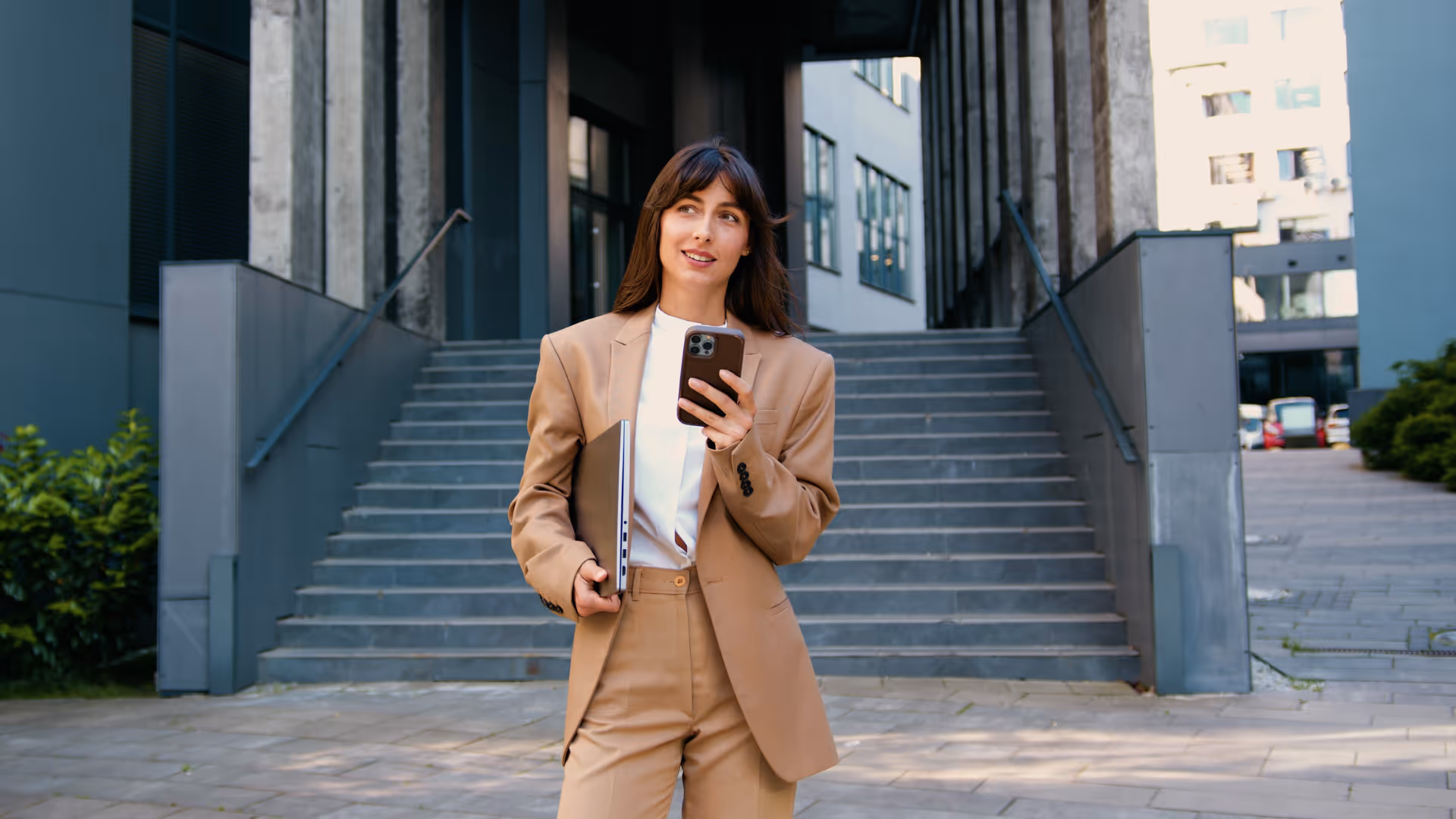 Woman in beige suit holding laptop and phone, standing in front of outdoor stairs at modern building.