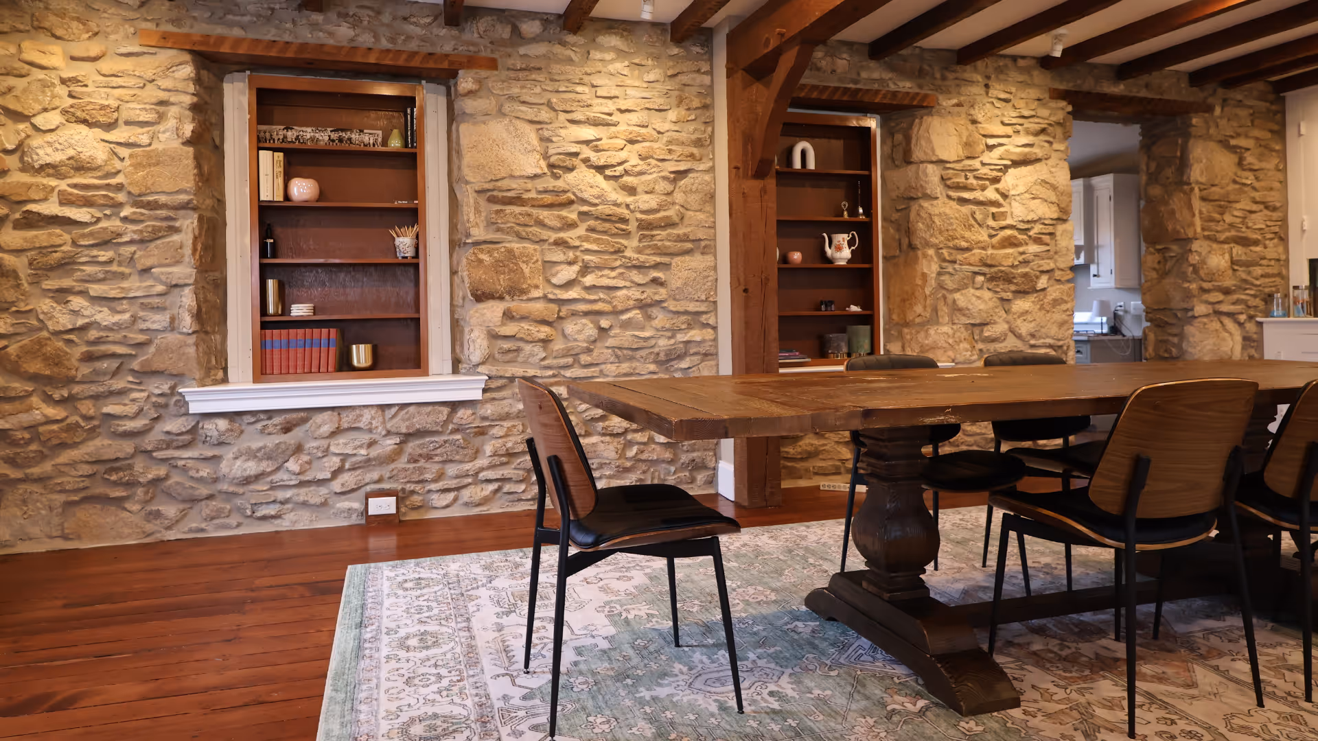 Dining area with wooden table, six modern chairs, stone walls, and built-in shelves with decor.