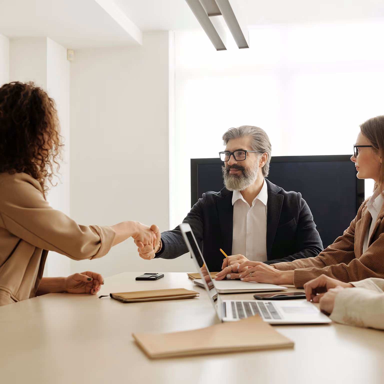 Man with gray beard shaking hands with woman across a meeting table with laptops and folders.