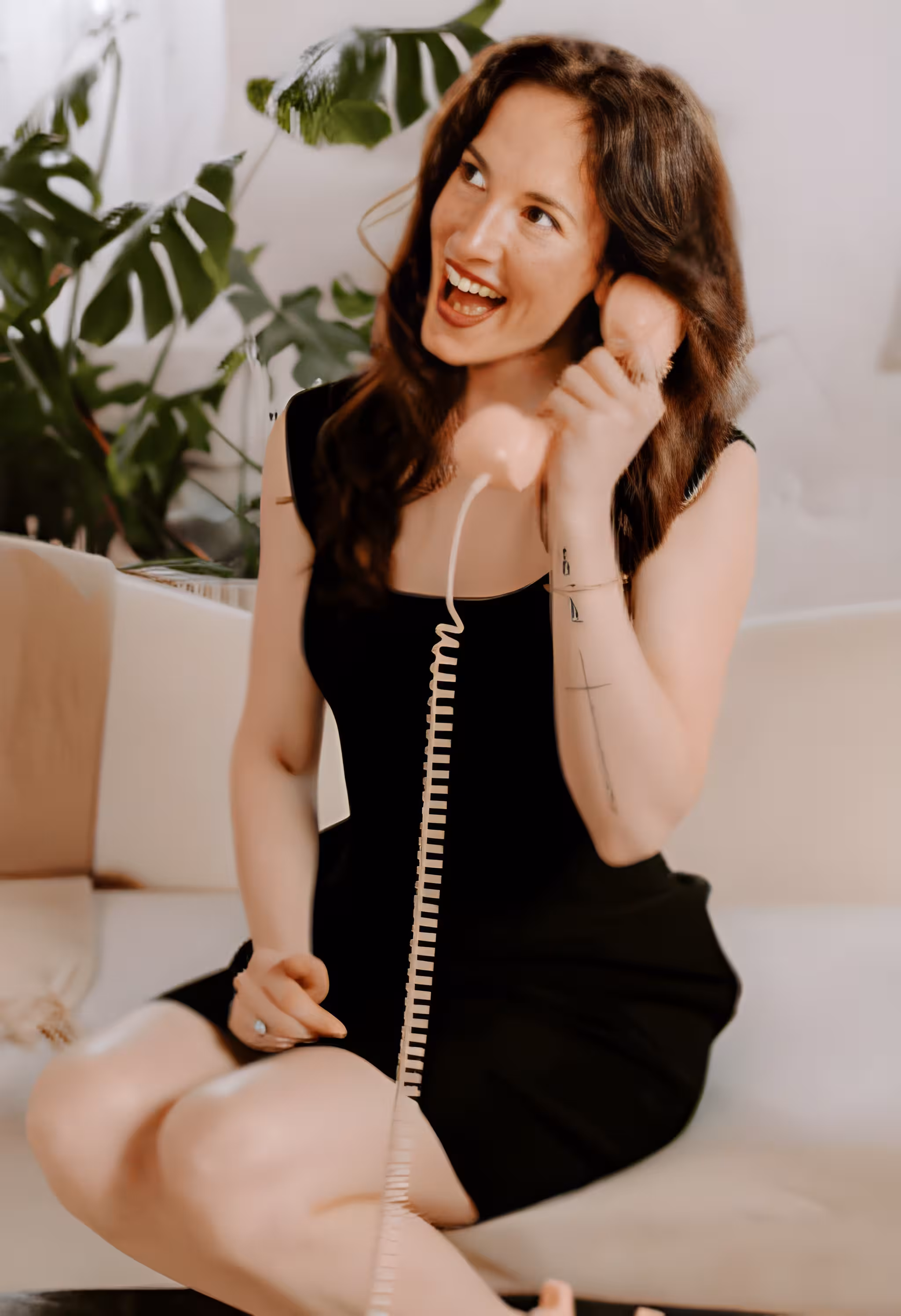 Smiling woman in a black dress sitting on a couch talking on a peach-colored corded phone.