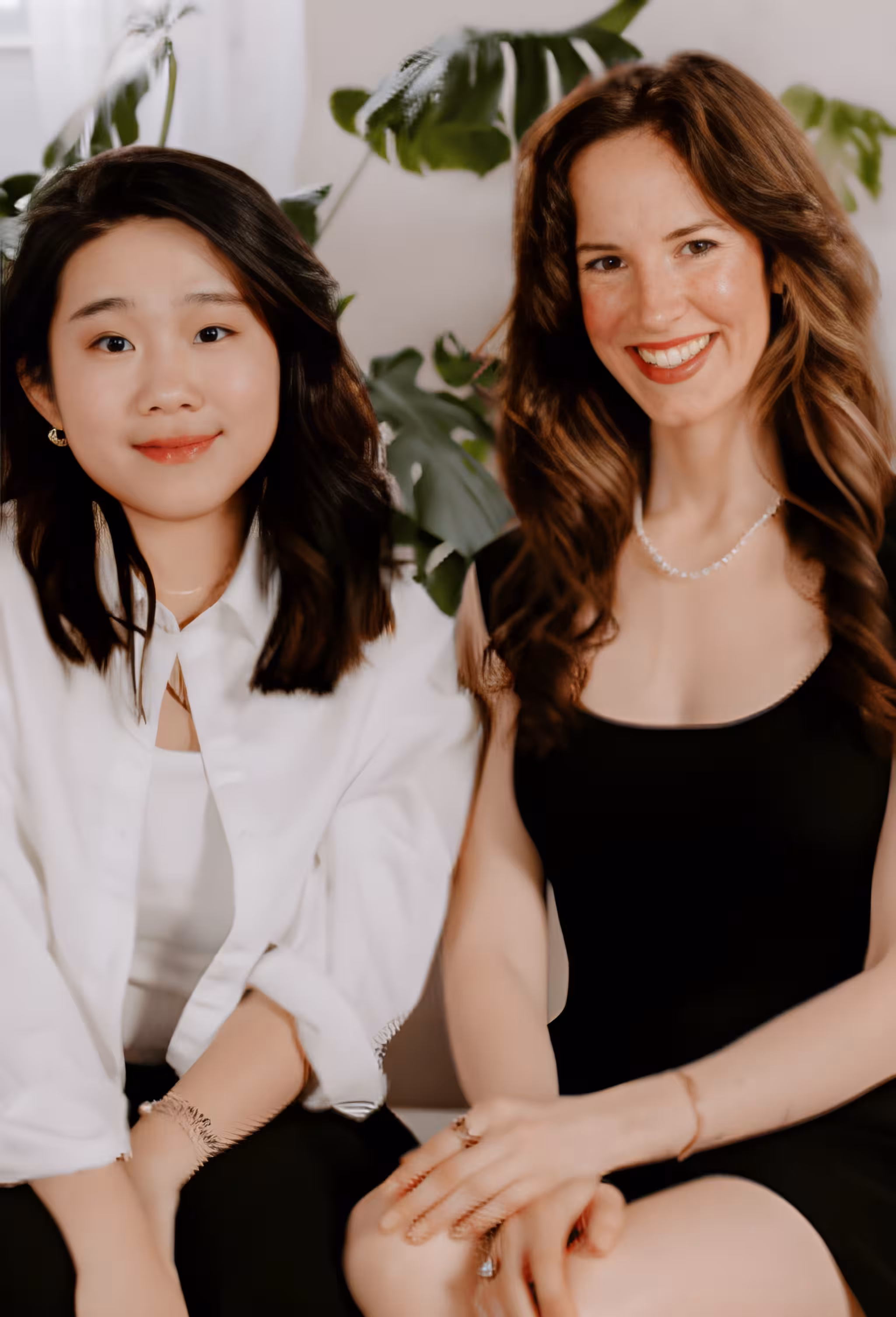 Two smiling women sitting side by side, one in white shirt and one in black dress with plants behind them.