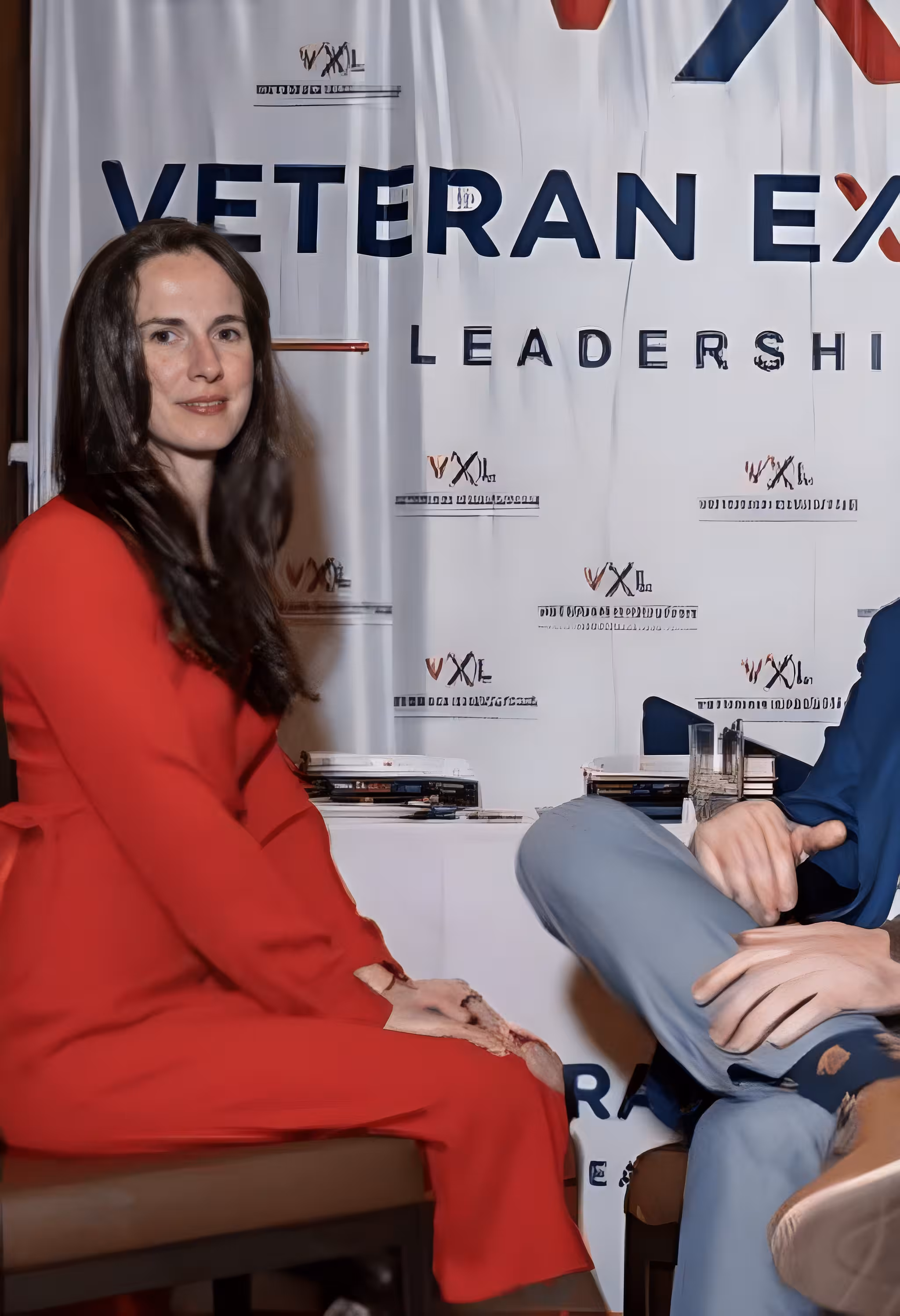 Woman in a red dress sitting next to a man, with a 'VETERAN EX LEADERSHIP' banner behind them.