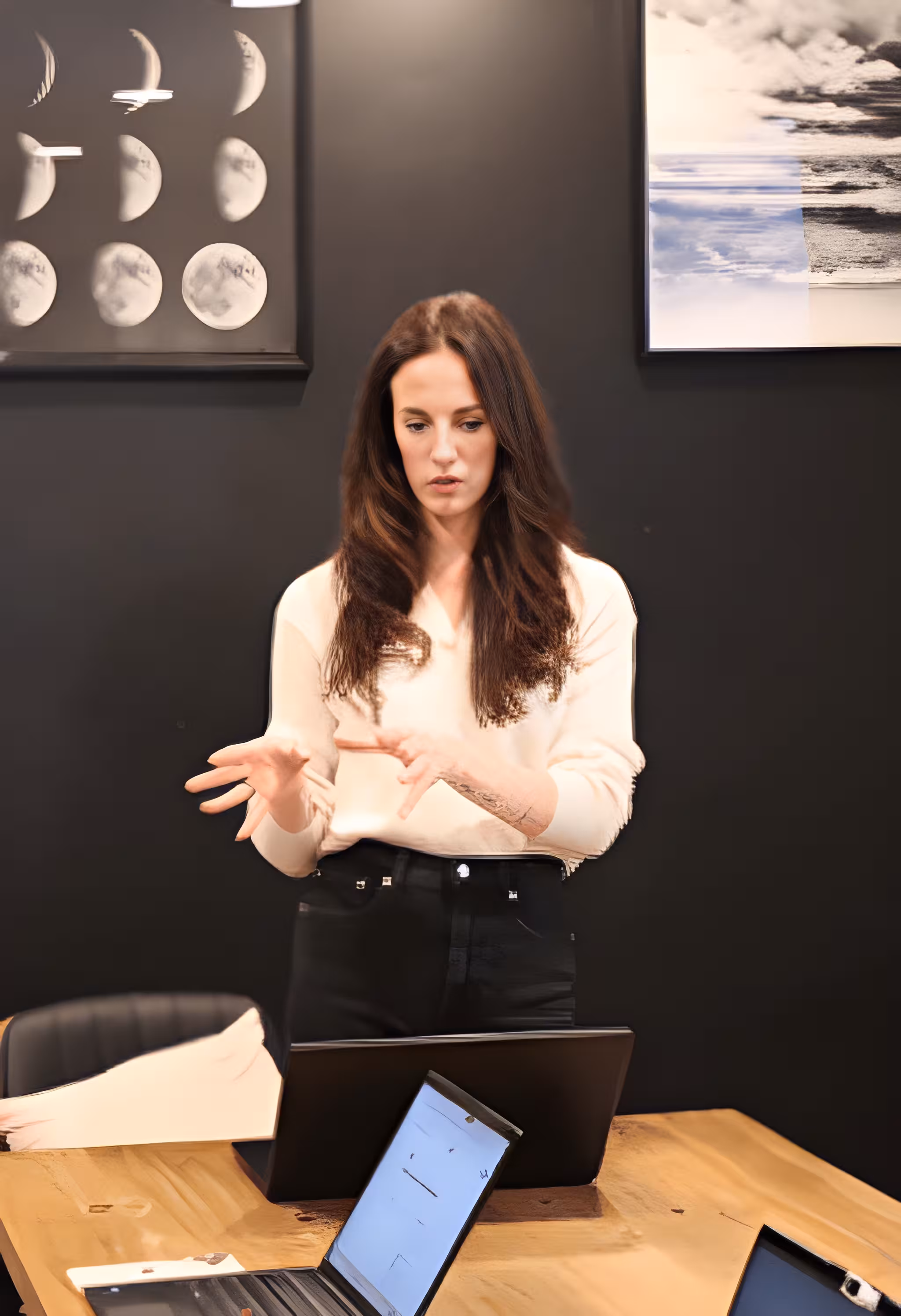 Woman with long brown hair gesturing in front of a laptop at a wooden table with moon phase art in background.