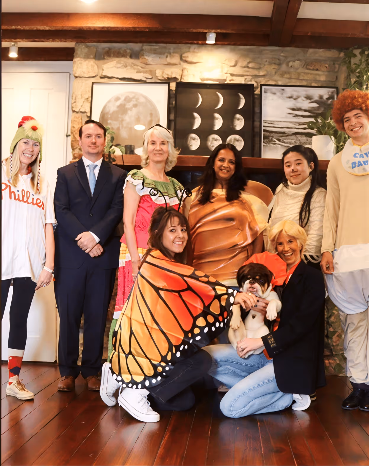 Group of eight people in costumes posing indoors, including butterfly, cracker, and baseball themes with a bulldog.