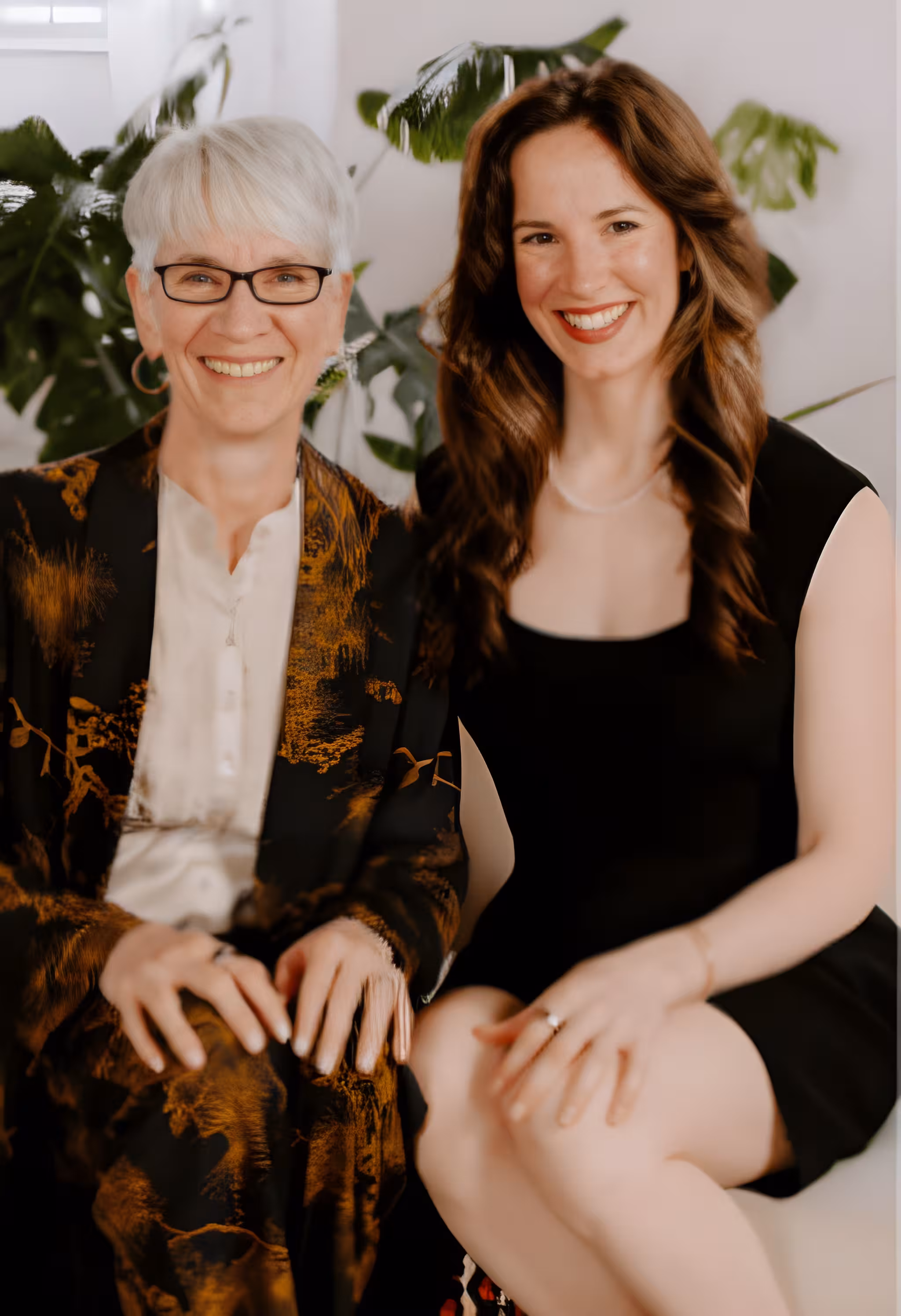 Two smiling women sitting side by side, one with short gray hair and glasses, the other with long brown hair.