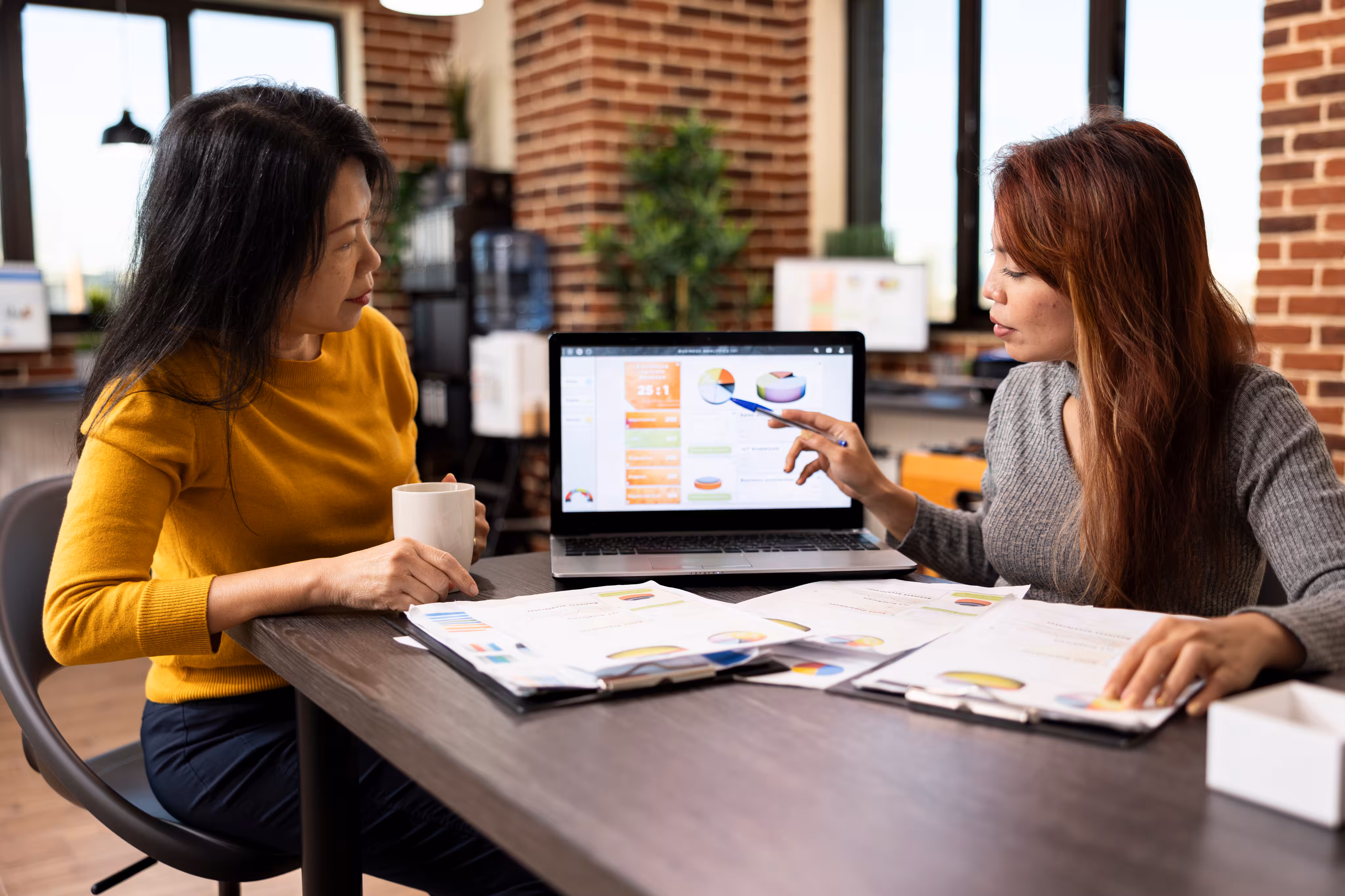 Two women analyzing charts and graphs on paper and laptop at a table in a modern office.