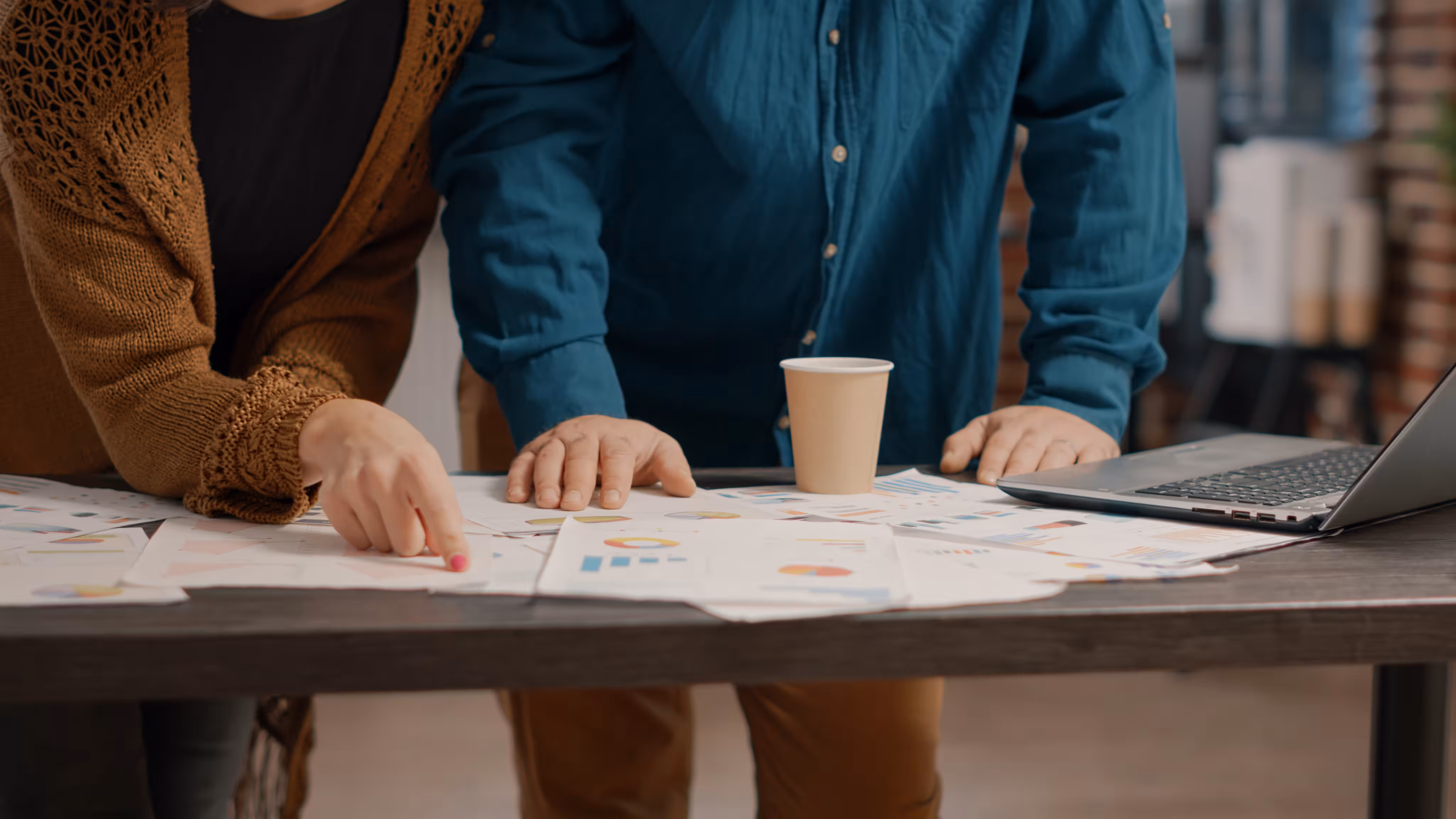 Two people reviewing charts and graphs spread over a table with a laptop and coffee cup.