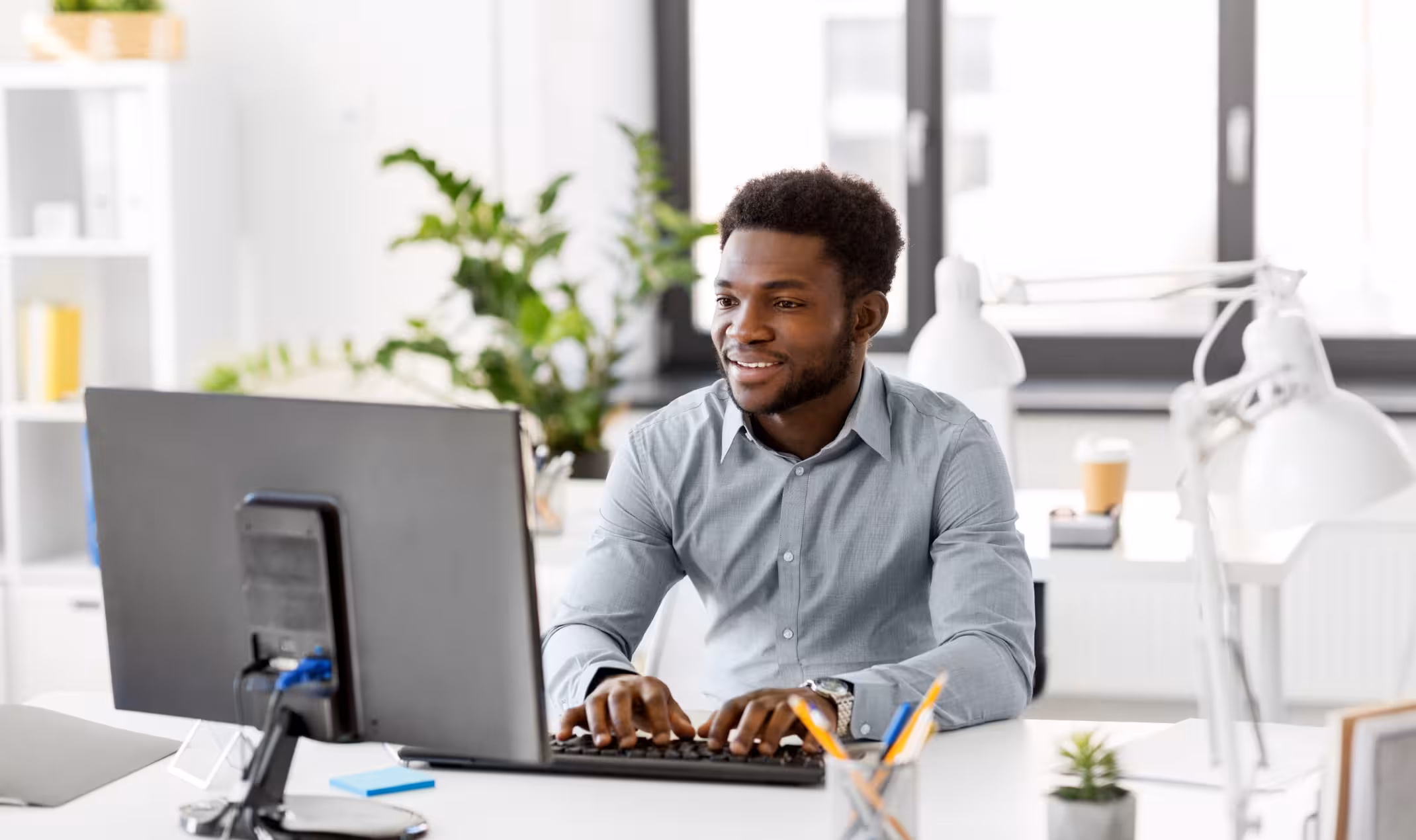 Smiling man in a blue shirt typing on a keyboard at a bright office desk with plants and lamps.