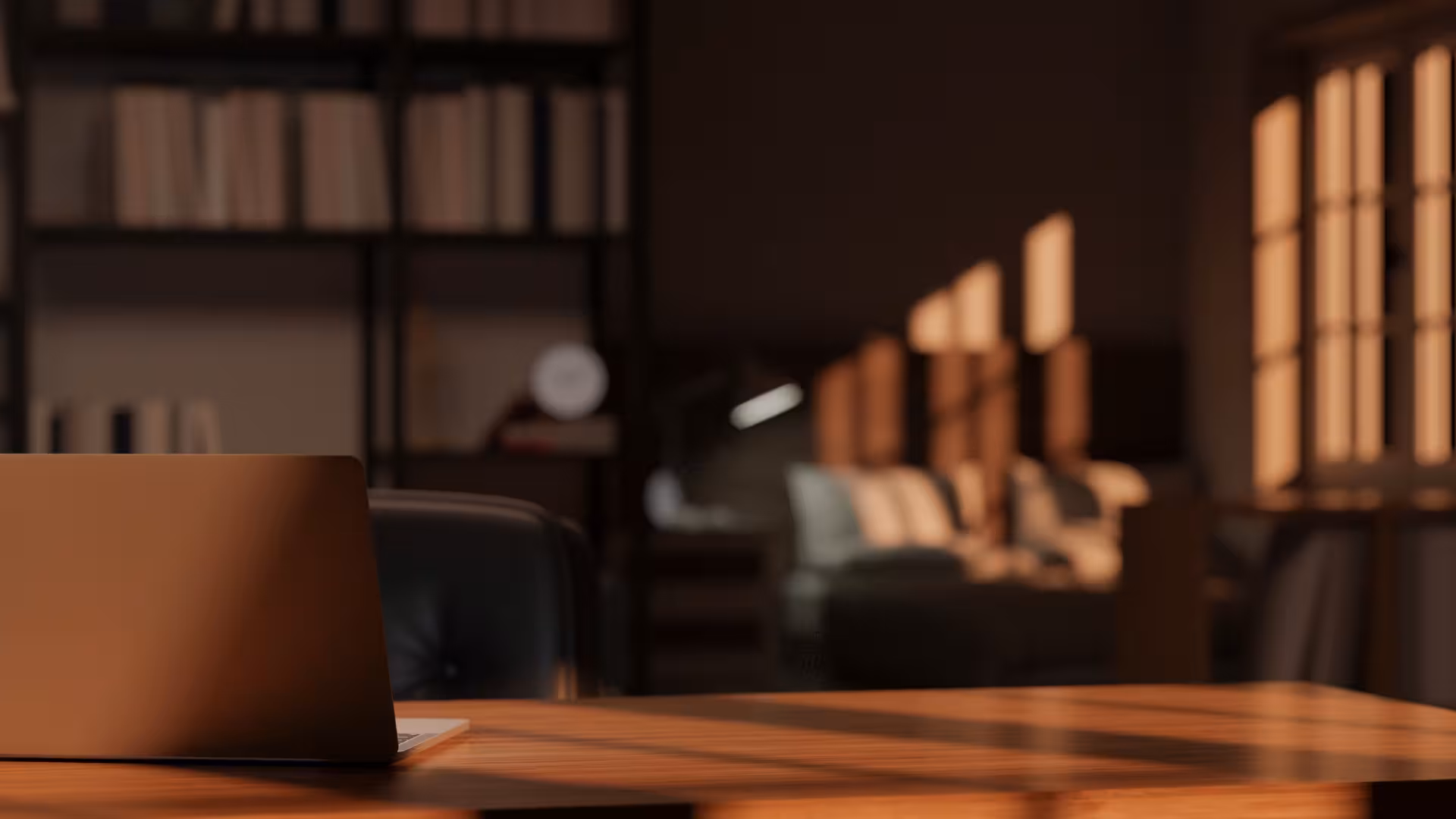 Closed laptop on wooden desk with warm sunlight in a cozy room with bookshelves and sofa.