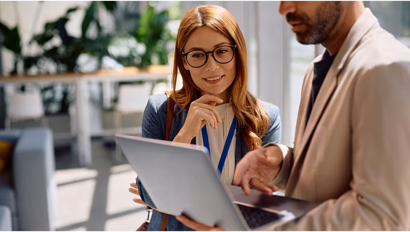 Two business colleagues discuss work on a laptop in a bright modern office.