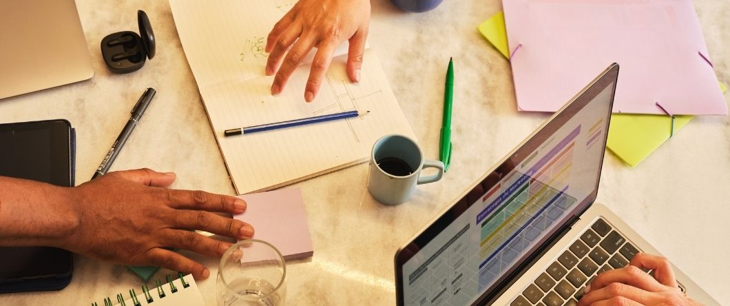 Two people working at a table with a laptop, notebook, pens, sticky notes, and a coffee cup.