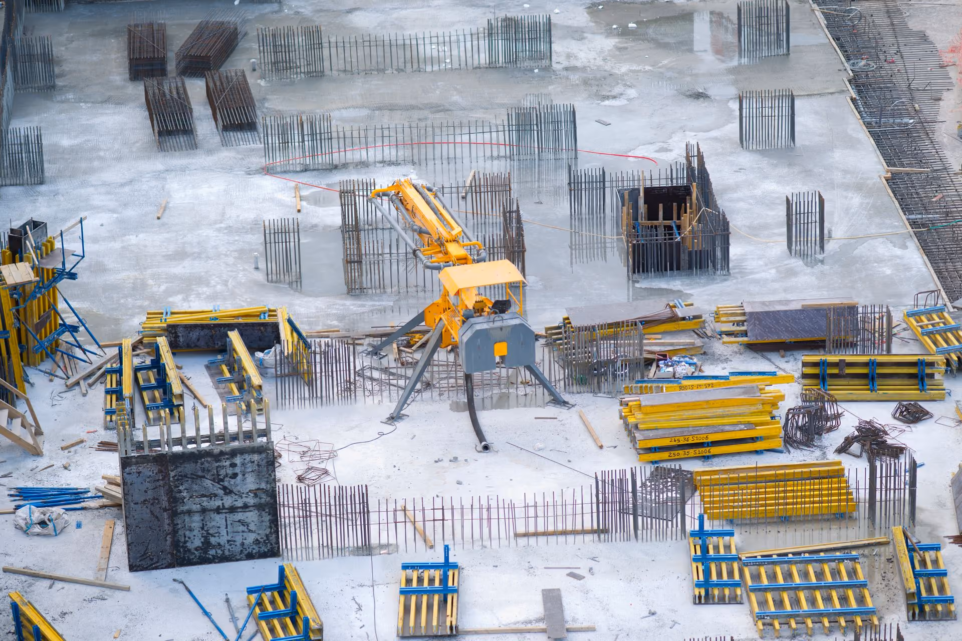 Construction site with yellow scaffolding, steel reinforcements, and a concrete pump machine on a wet slab.