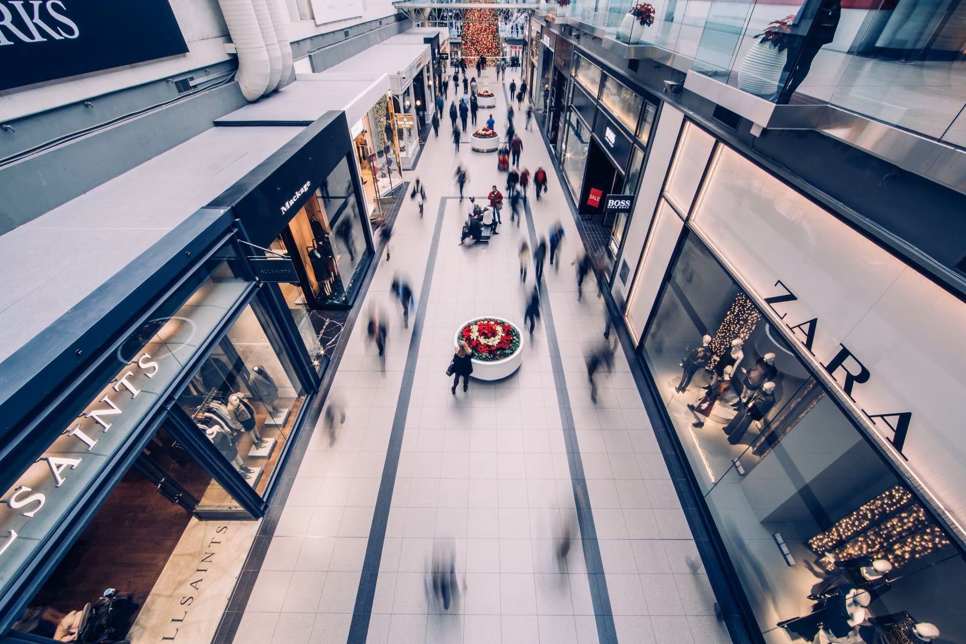 Blurred shoppers walking in a mall corridor lined with AllSaints and Zara stores with holiday decor.
