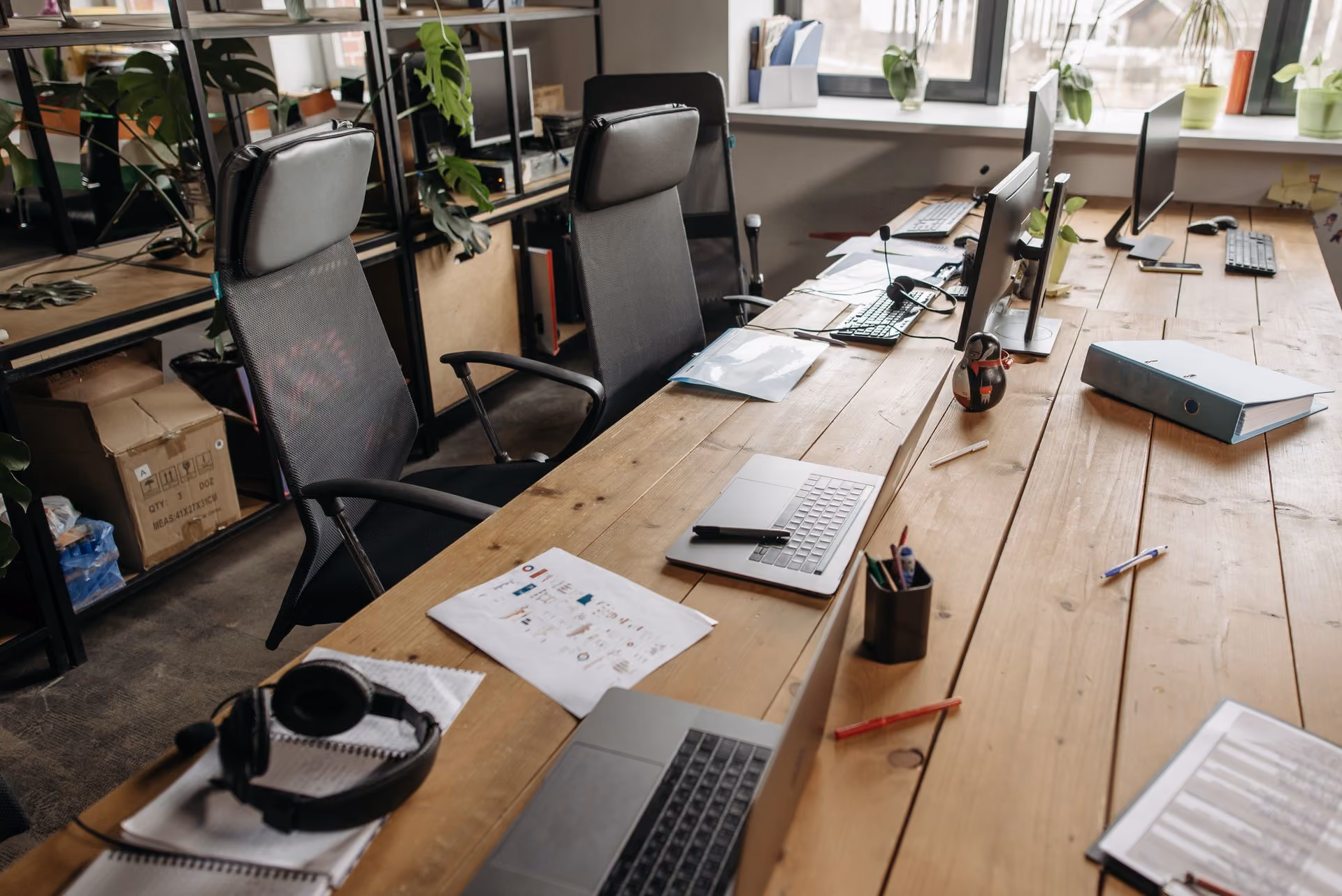 Wooden office desk with laptops, monitors, papers, headphones, and office chairs in a workspace.