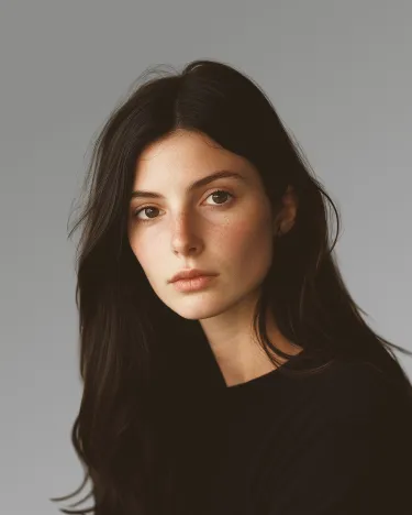 Portrait of a young woman with long dark hair wearing a black top against a gray background.