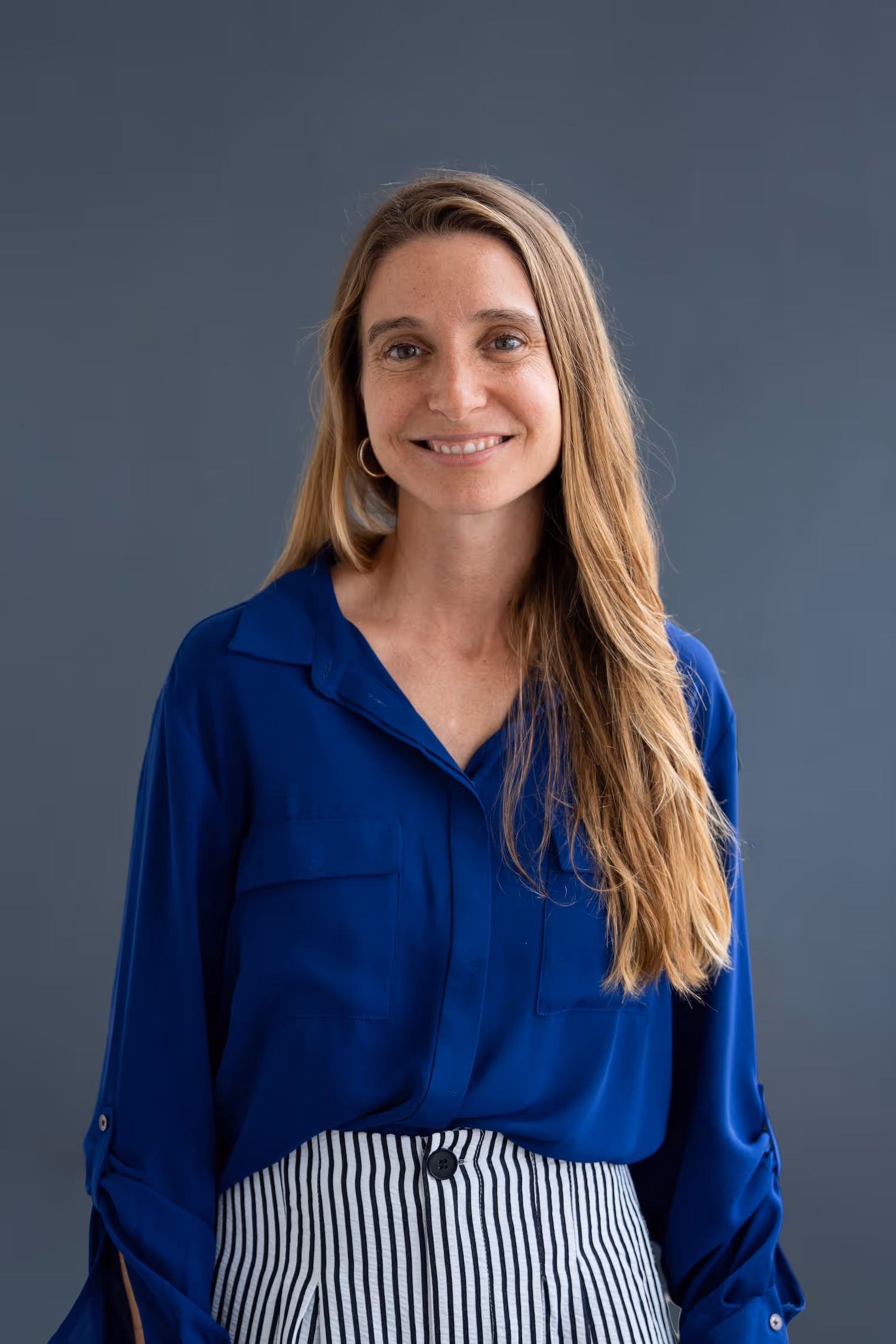Smiling woman with long blonde hair wearing blue blouse and striped pants against gray background.
