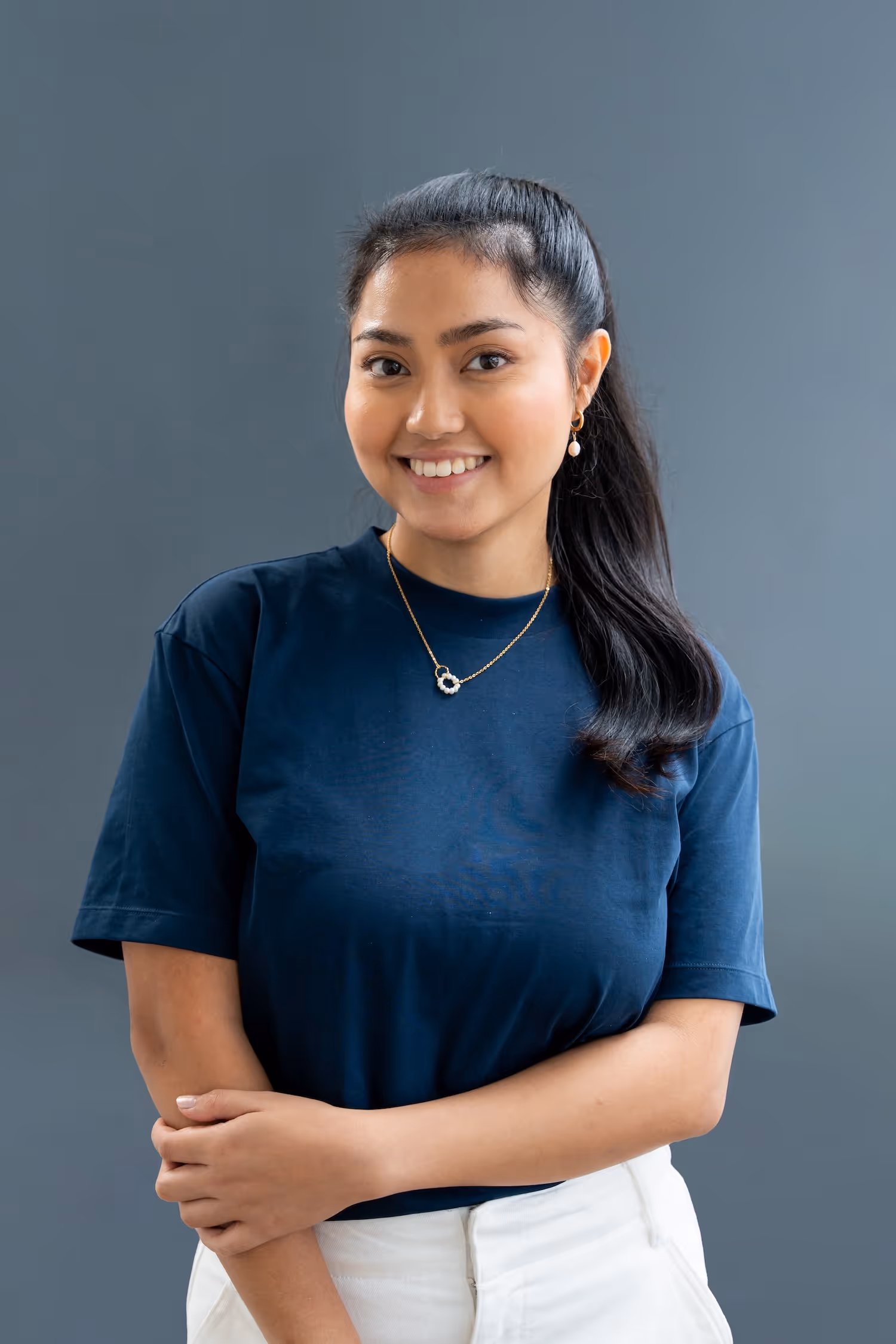 Smiling woman with black hair in ponytail wearing navy shirt, white pants, gold necklace, and earrings.