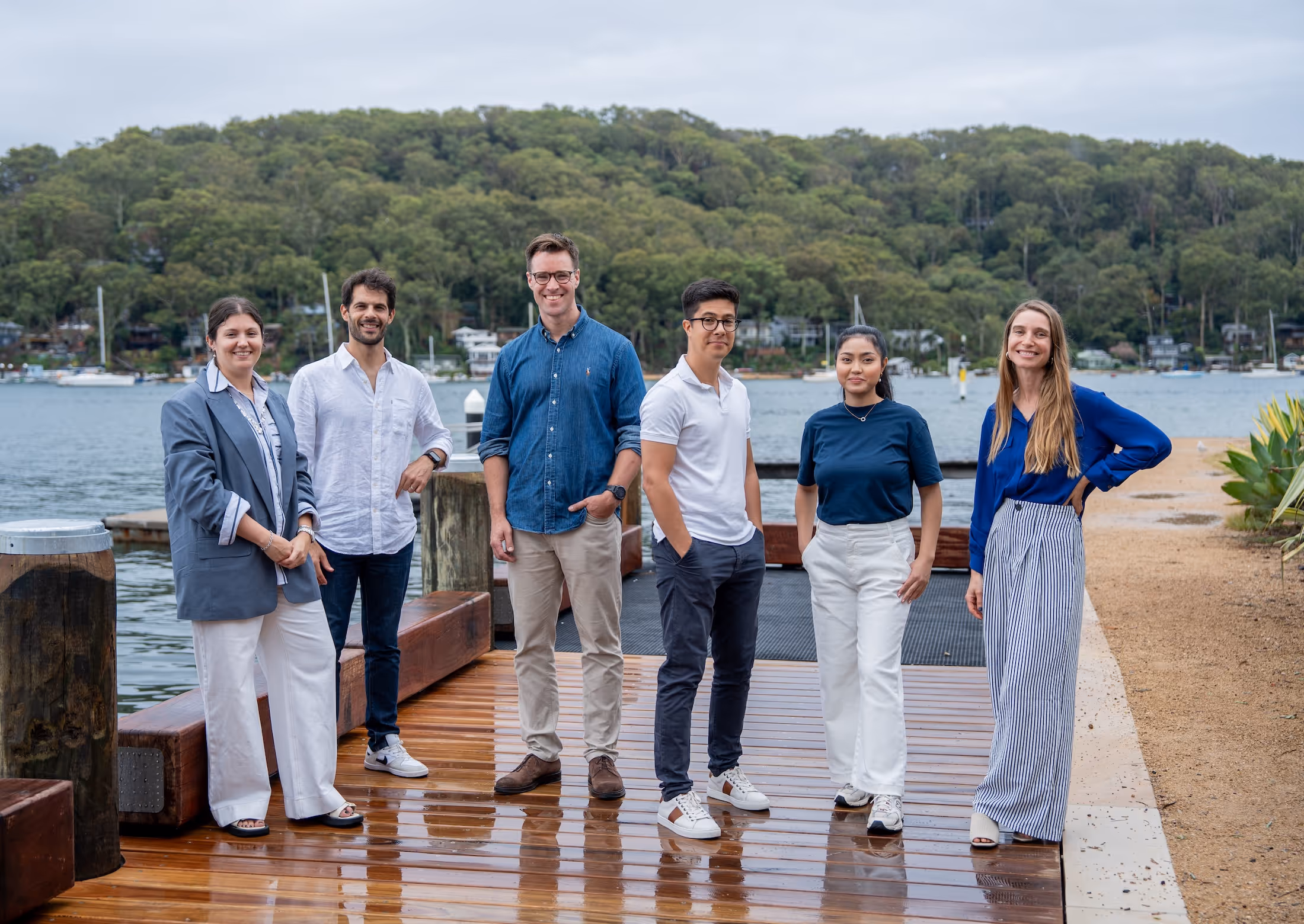 Six people standing on a wooden dock by water with trees and boats in the background.