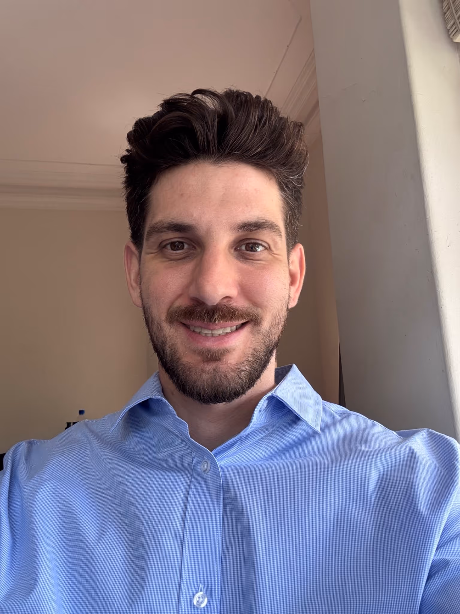 Smiling man with dark hair and beard wearing a light blue collared shirt indoors near a window.