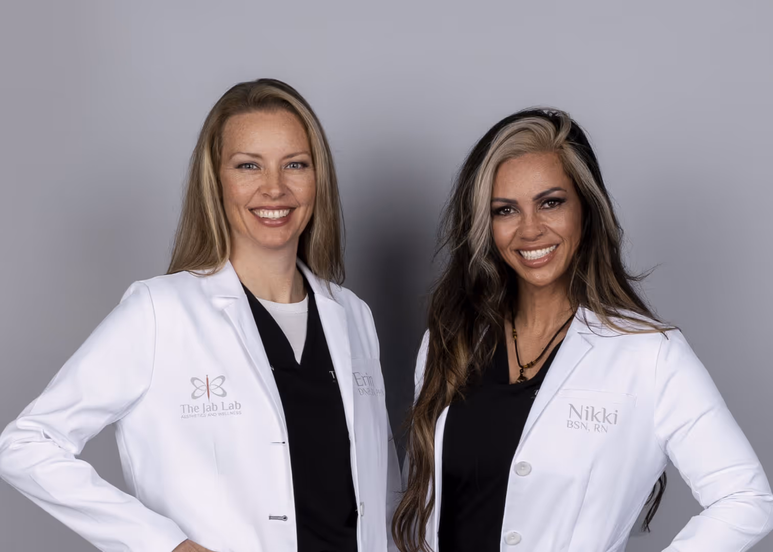 Two female medical professionals in white lab coats smiling and posing against a gray background.