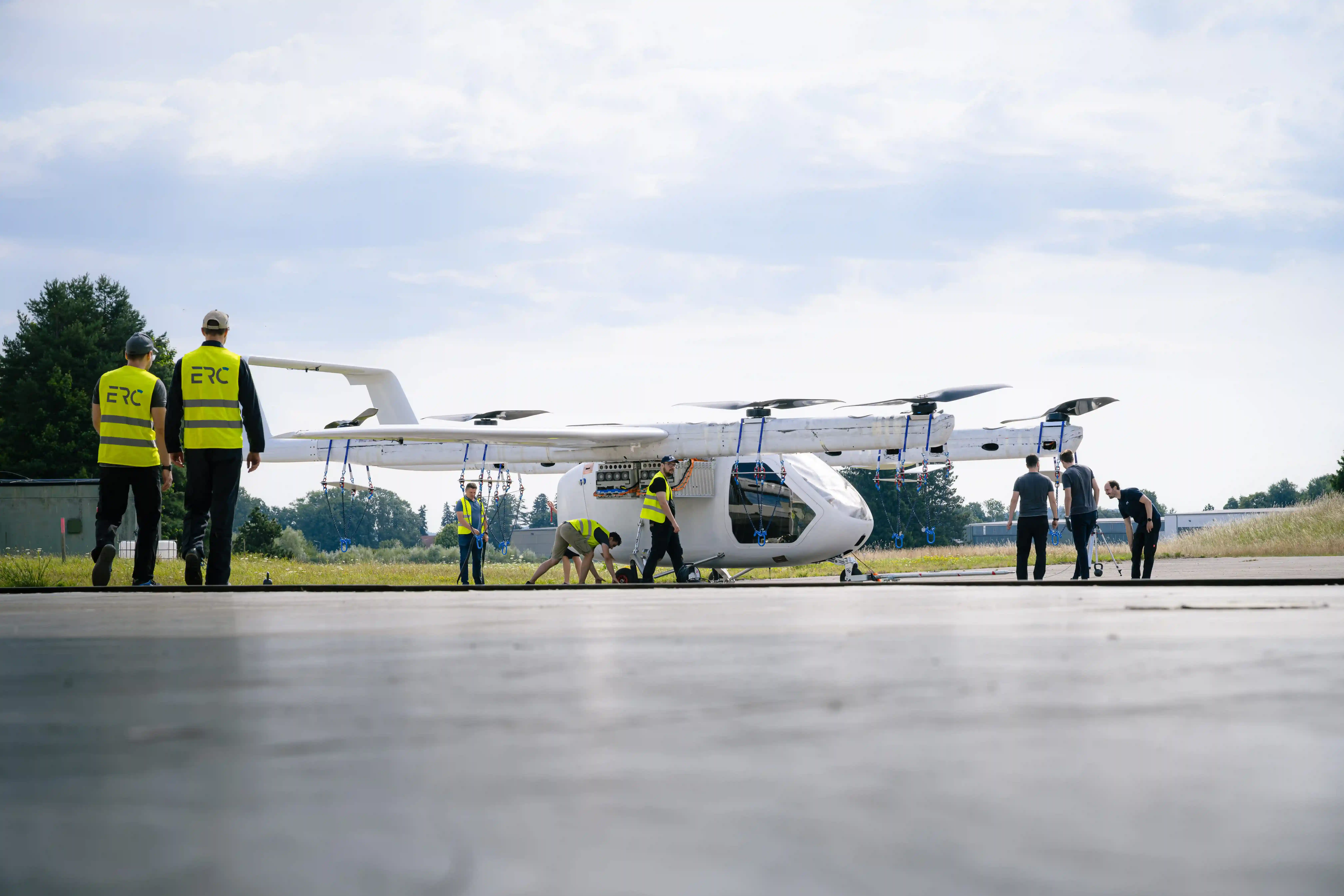 People wearing yellow vests inspecting a white electric vertical takeoff and landing aircraft outdoors.