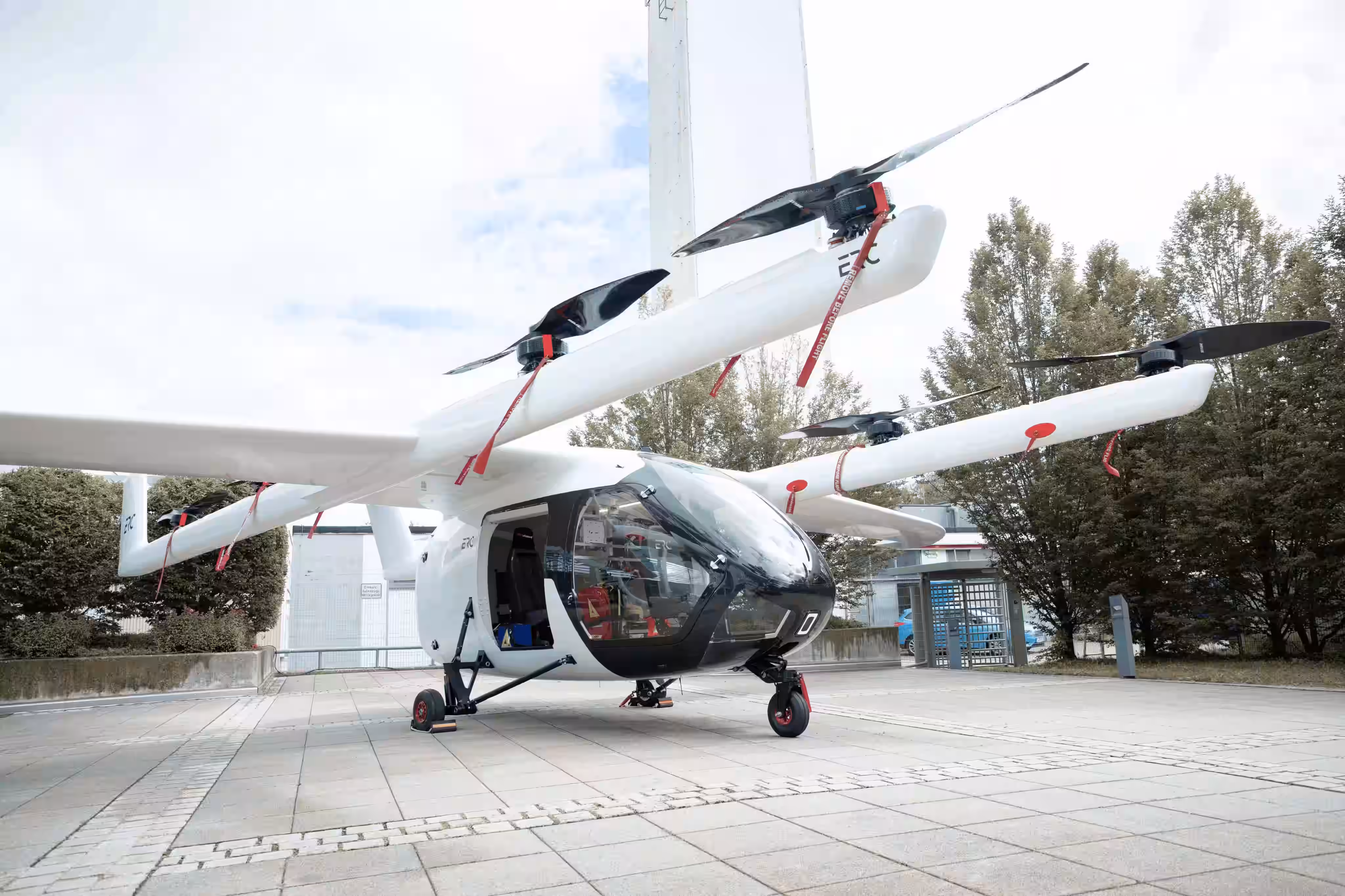 White electric vertical takeoff and landing aircraft with open door on paved area near trees.