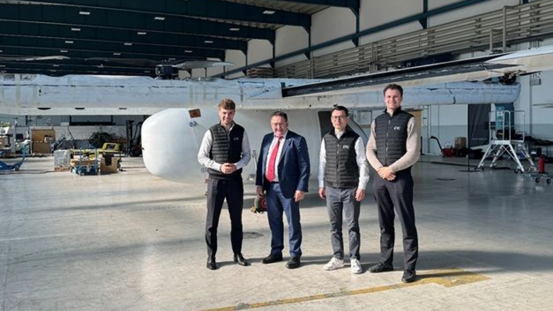 4 people in formal clothing standing side by side in front of the ERC Charlie C700 aircraft in a hangar.