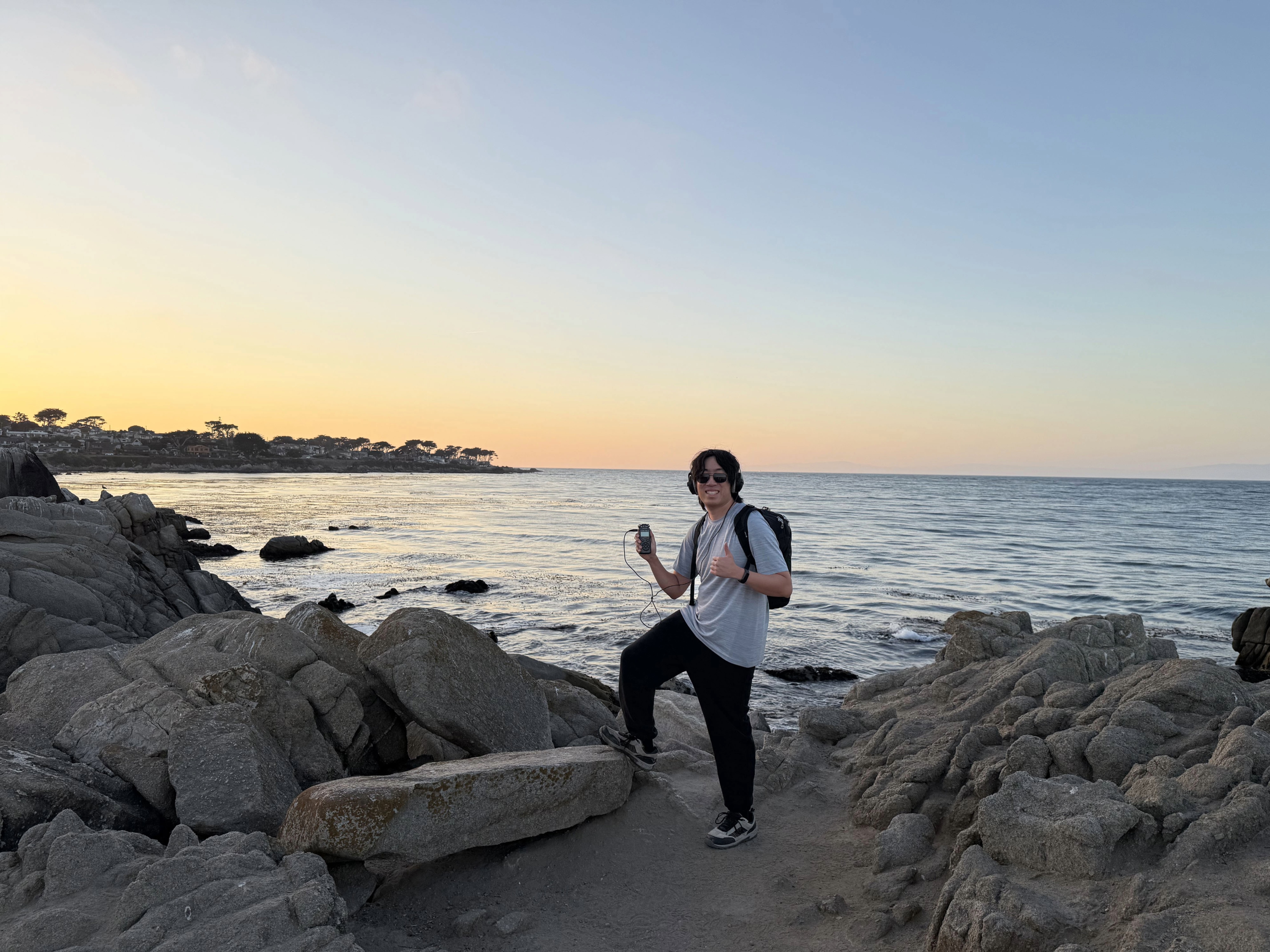 A man with sunglasses, a backpack, and a handheld audio recording device poses for a picture by the ocean at Lovers Point Park at around golden hour