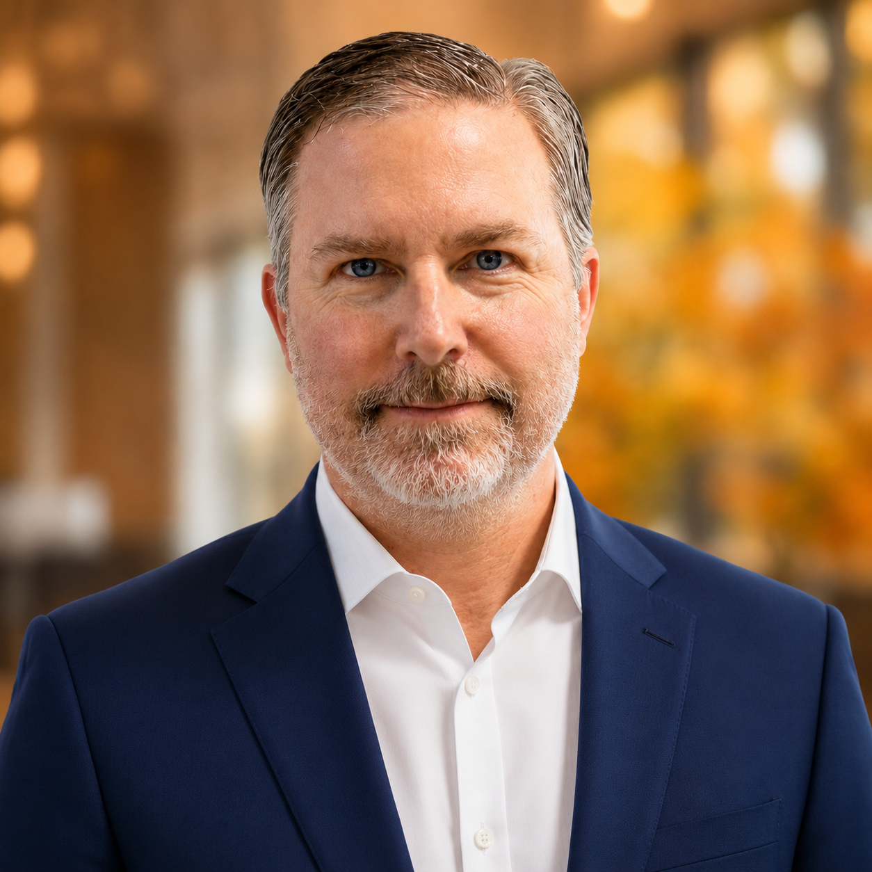 Portrait of a man, short salt-and-pepper hair, and beard, wearing a navy blue suit jacket and white shirt, against a blurred warm-toned background.