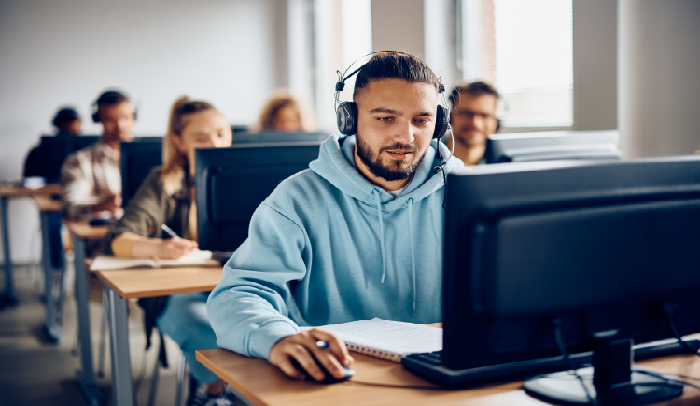 Man in a blue hoodie and headset working on a desktop computer in a classroom with other students.