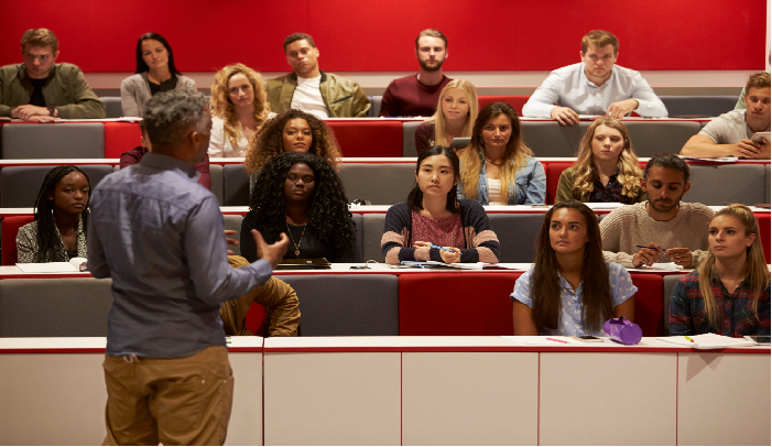 Diverse university students attentively listening to an instructor in a lecture hall with red and gray seats.