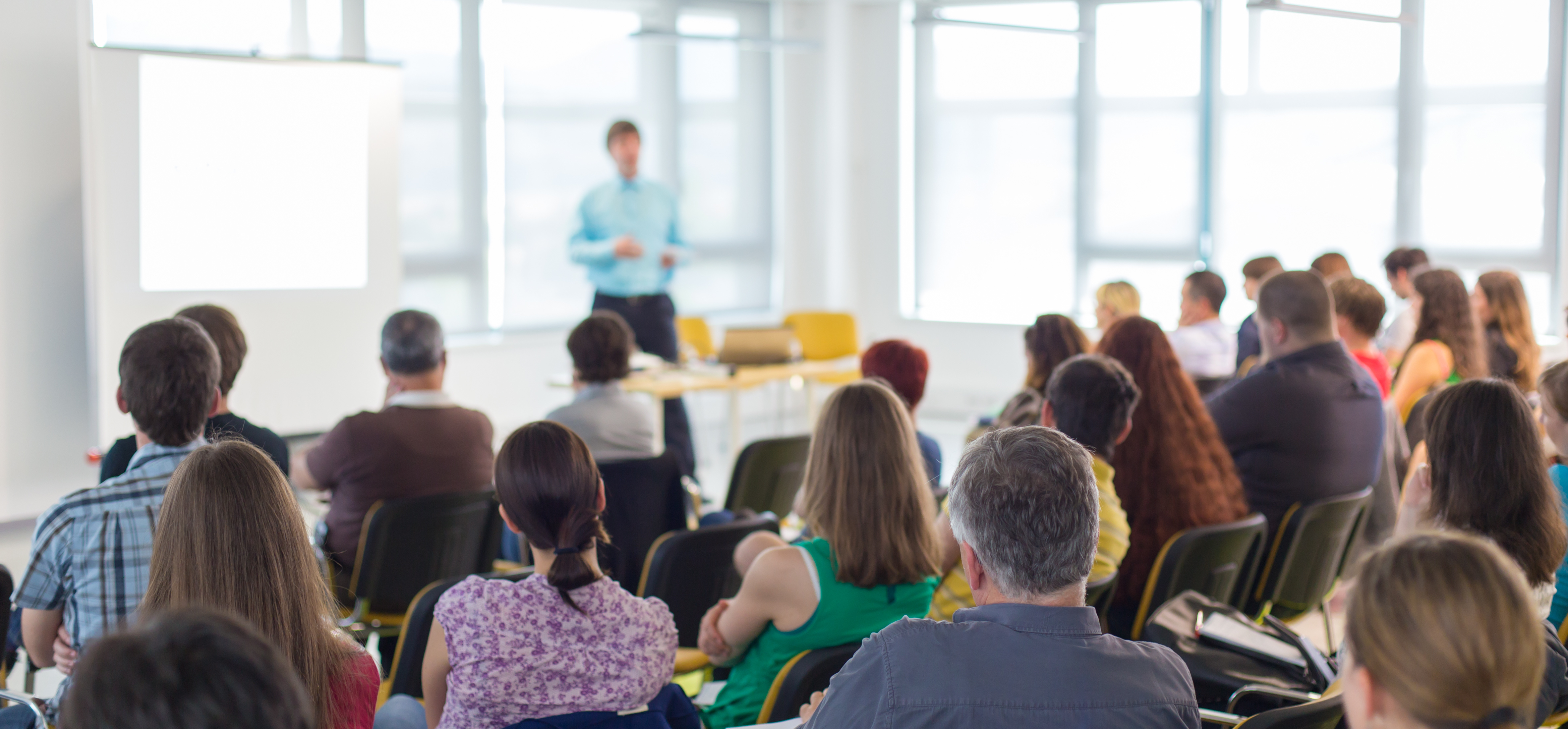 Audience seated in a bright room listening to a presenter near a blank white screen.